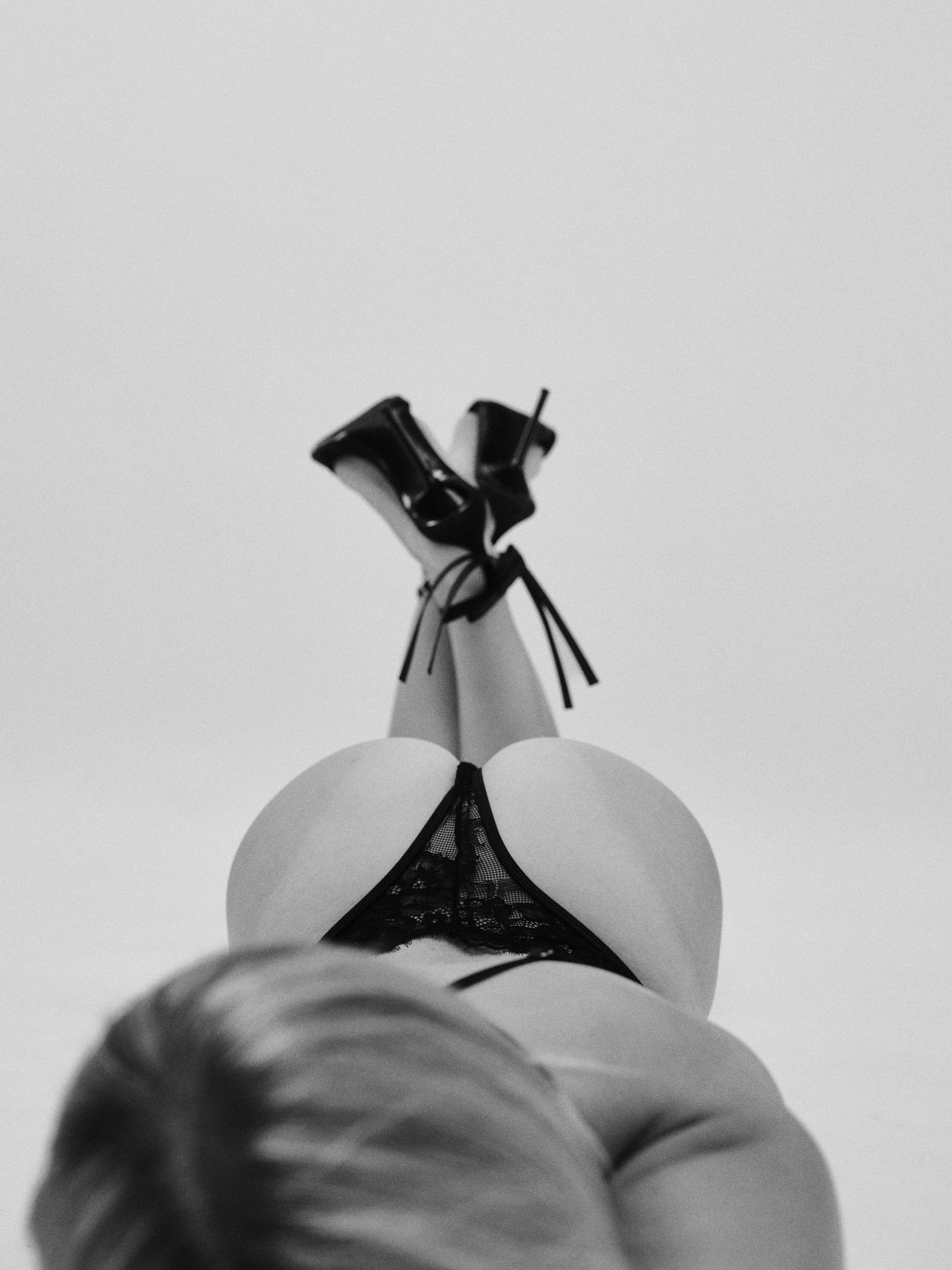 Black and white photo of a person in lace lingerie and heels, legs raised overhead against a light background.