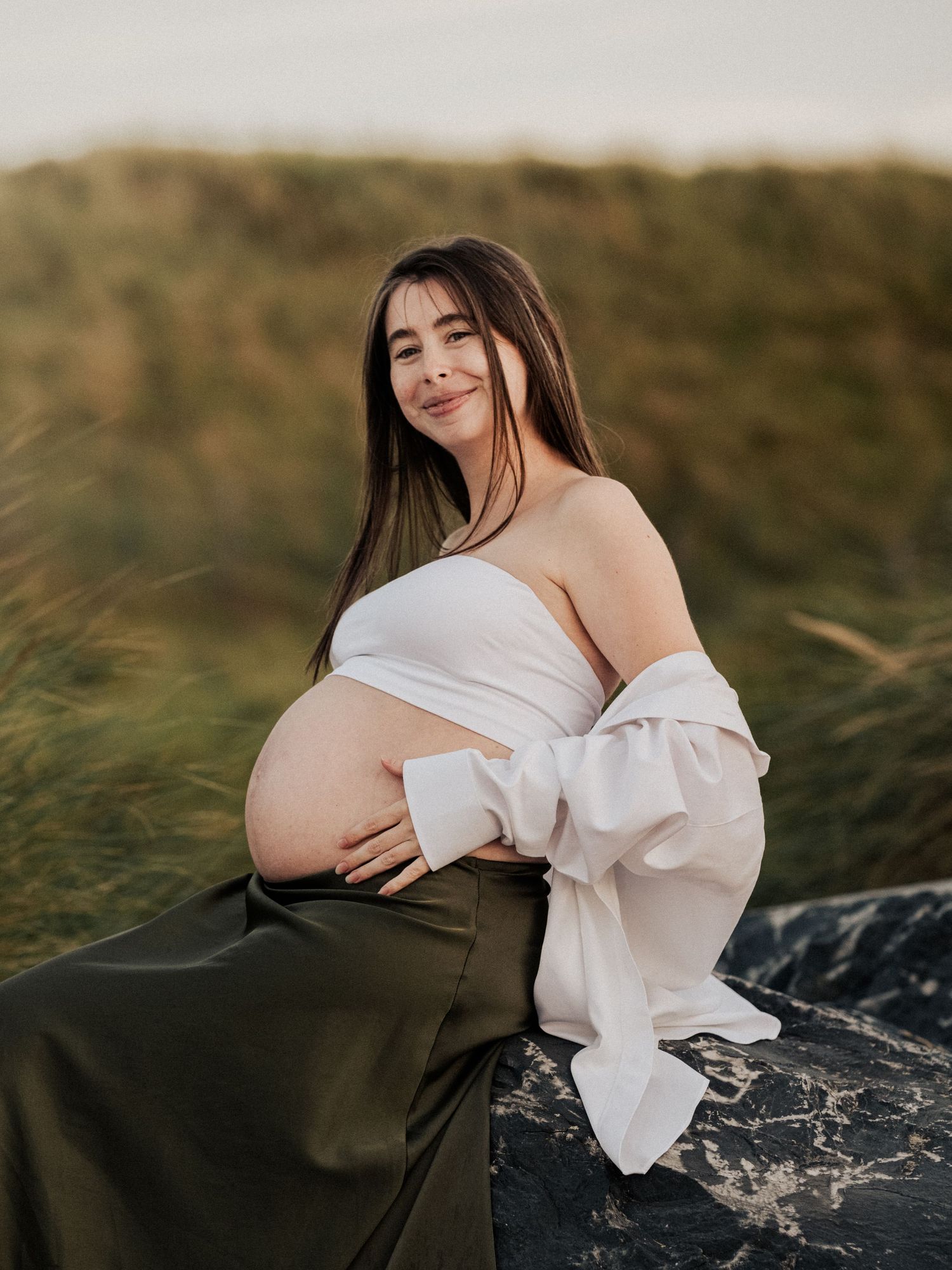 A maternity photoshoot in a grassy field at sunset with a white off-shoulder top and flowing dark skirt.