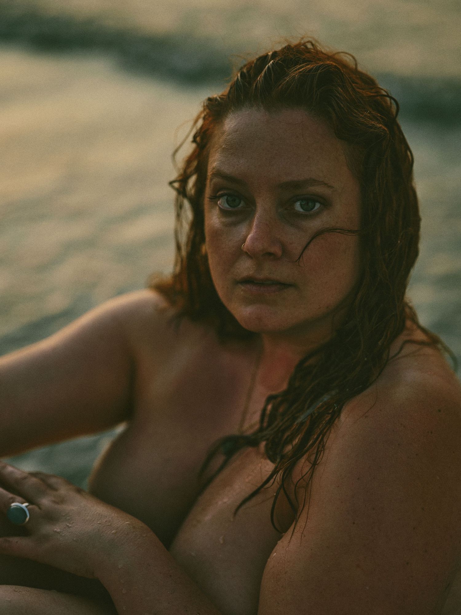 A wet-haired woman sits by the water's edge at golden hour, gazing directly at the camera.