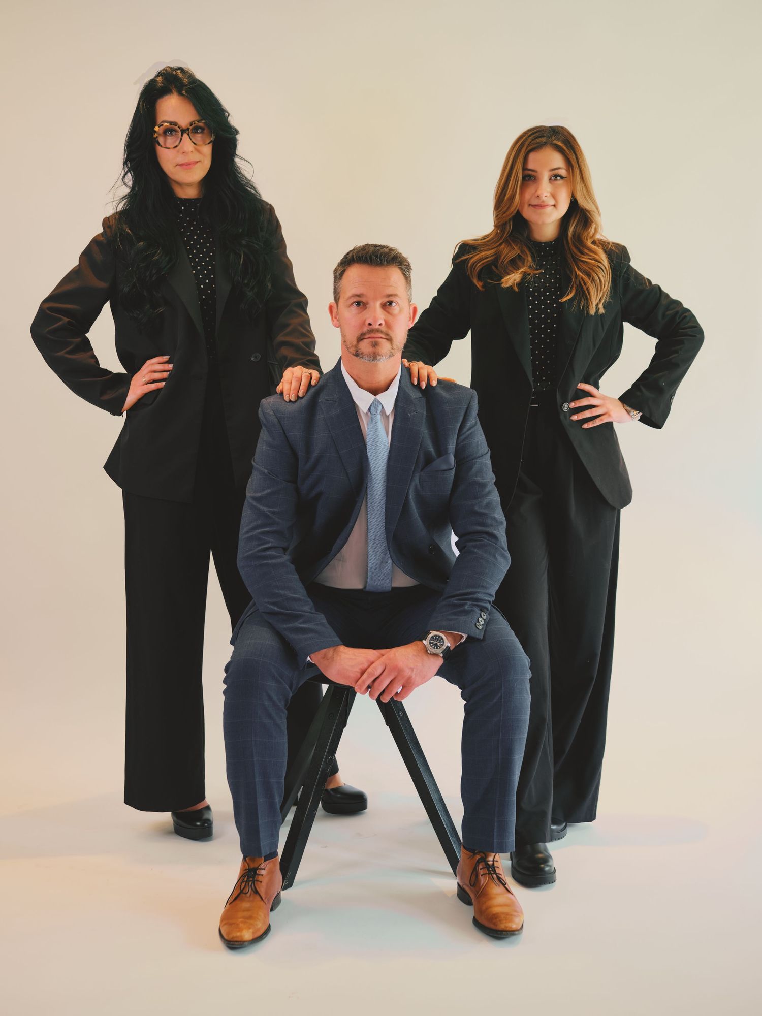 Three business professionals in formal attire pose confidently, one man seated and two women standing, against a white background.