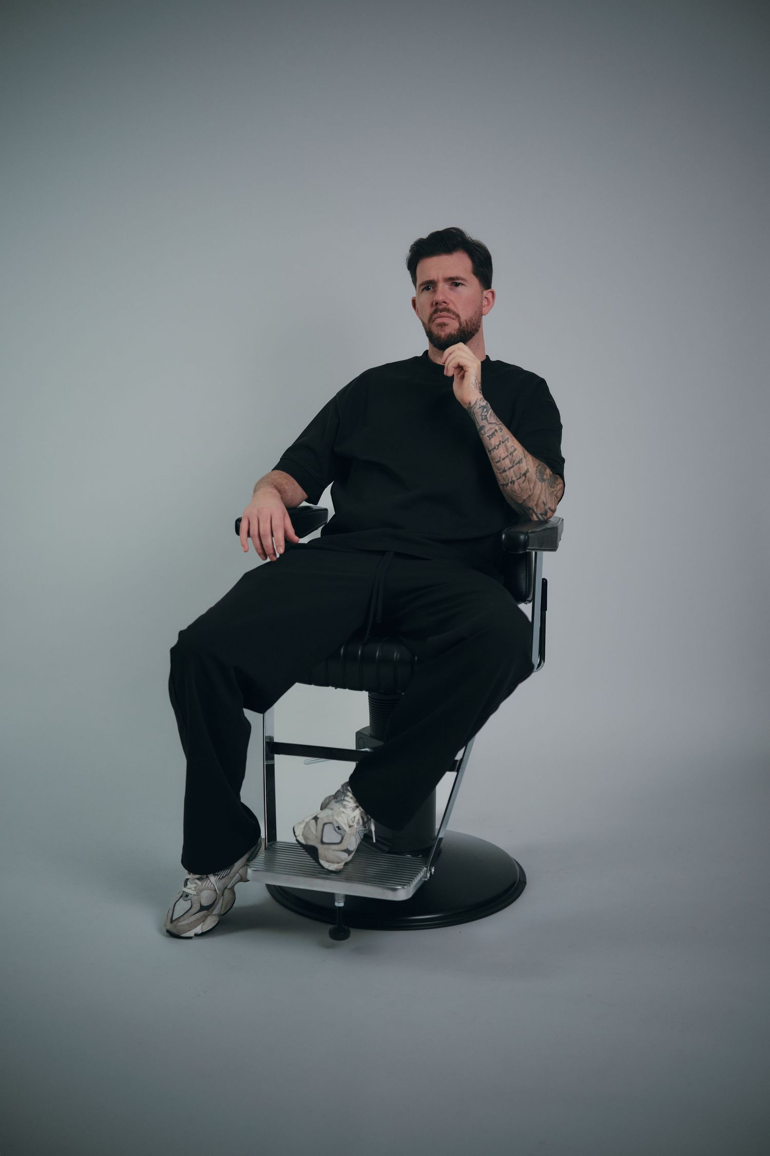 A person in black clothing sits in various poses on a chair against a gray studio backdrop.