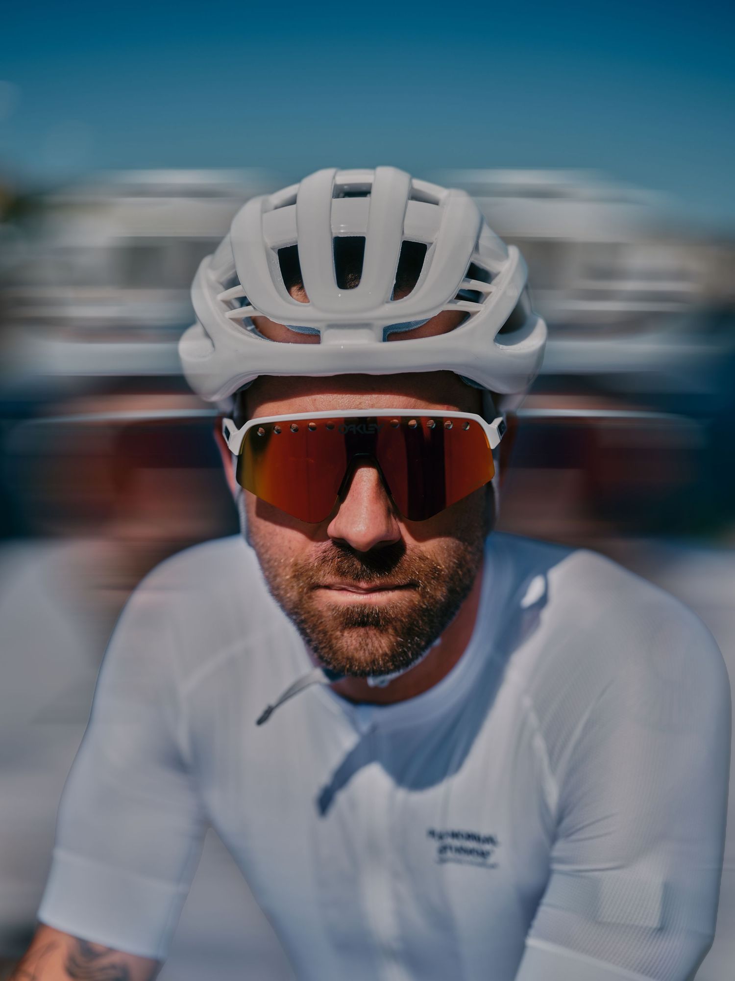 A male cyclist in a white helmet and red sport sunglasses rides at speed, wearing a white jersey on a sunny day.
