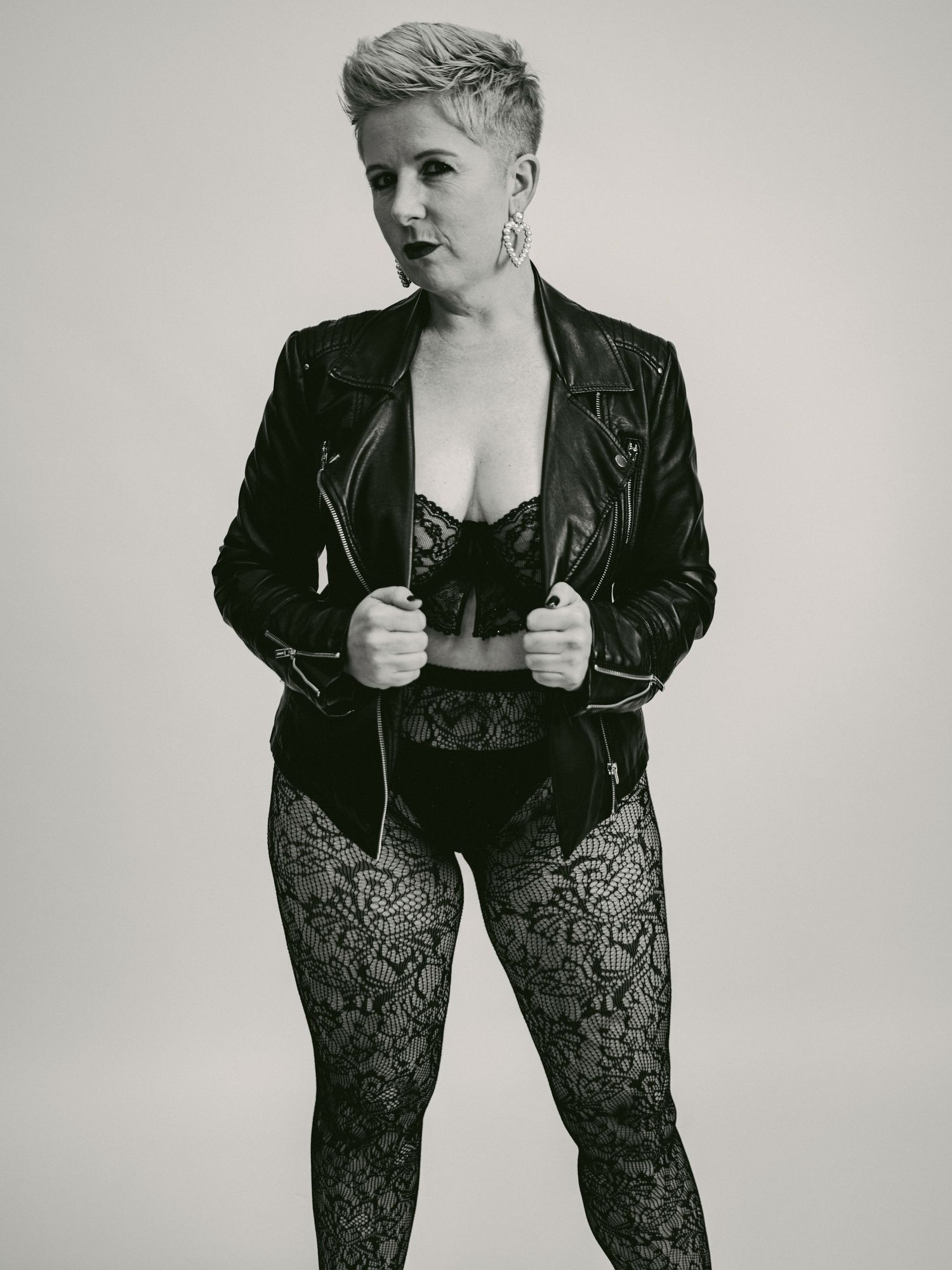 Black and white portrait of someone wearing a leather jacket and lace pants in an edgy fashion pose against a plain backdrop.
