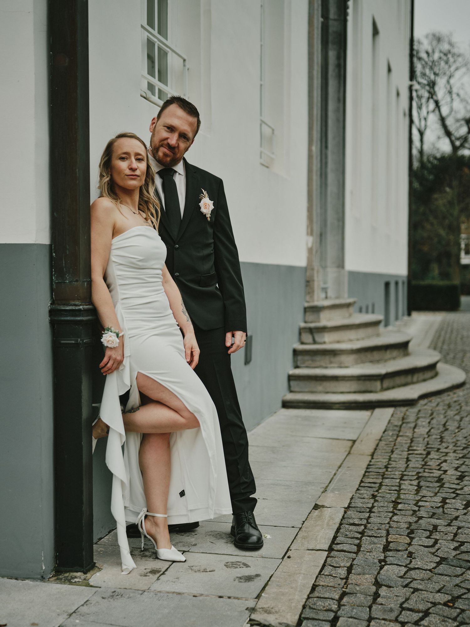 A couple poses together against a white building wall on a cobblestone street during their wedding photoshoot.