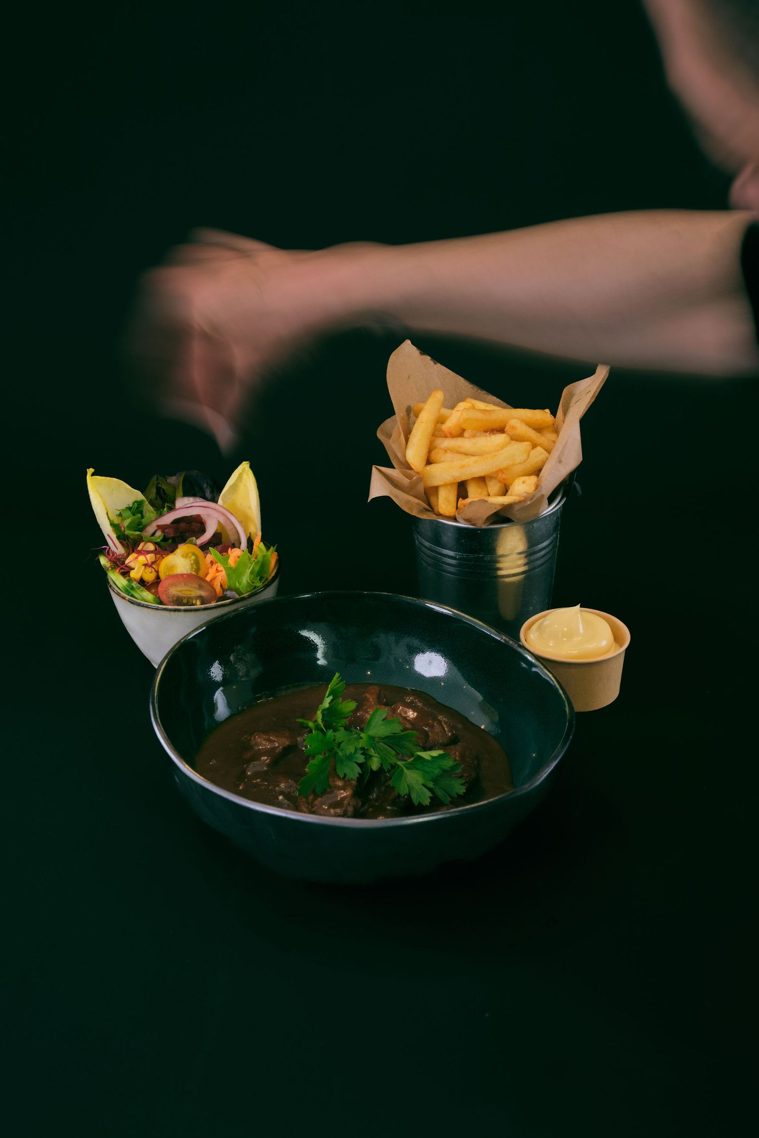A dark bowl of beef stew with parsley, served with fresh salad, golden french fries in a bucket, and dipping sauce.