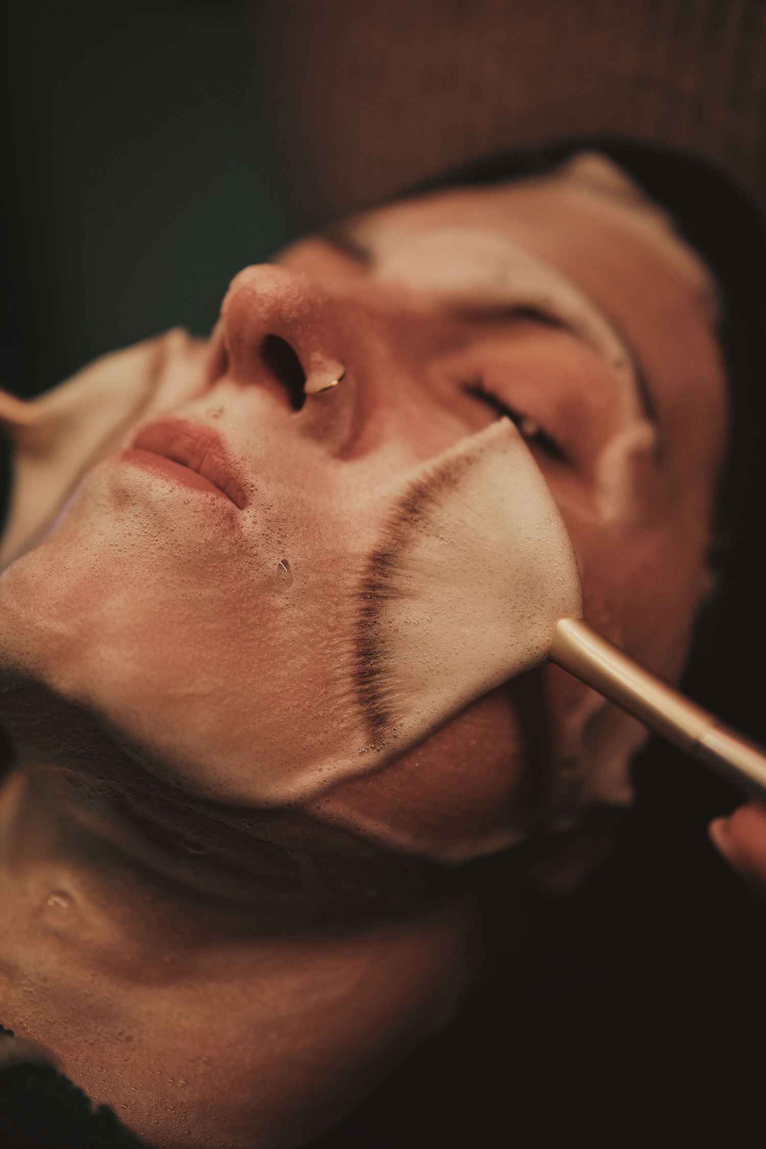 Esthetician applying a facial mask with a brush on a client's face during a skincare treatment.