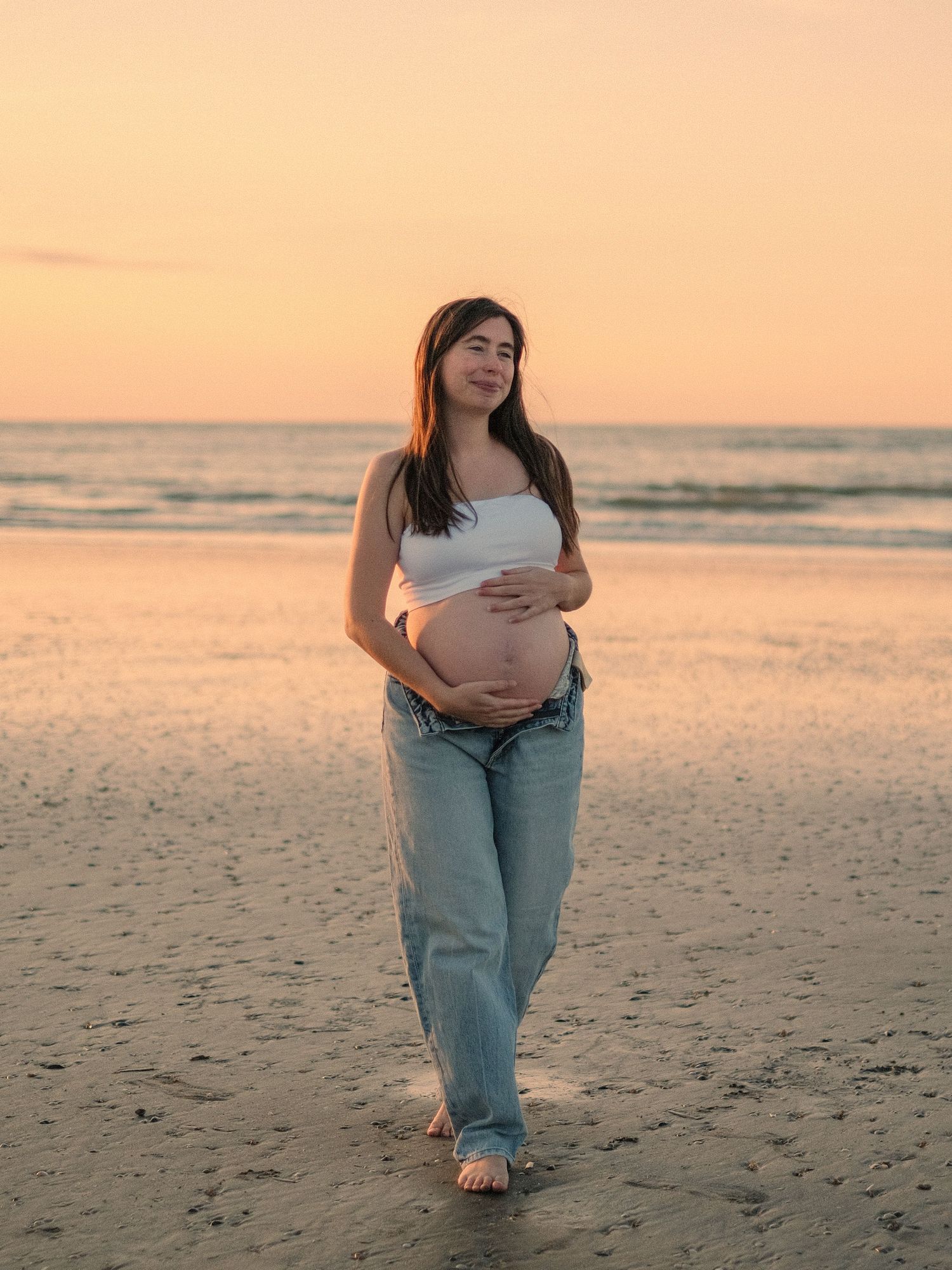 A person in light blue jeans walks barefoot on a sandy beach at sunset creating a peaceful coastal scene.