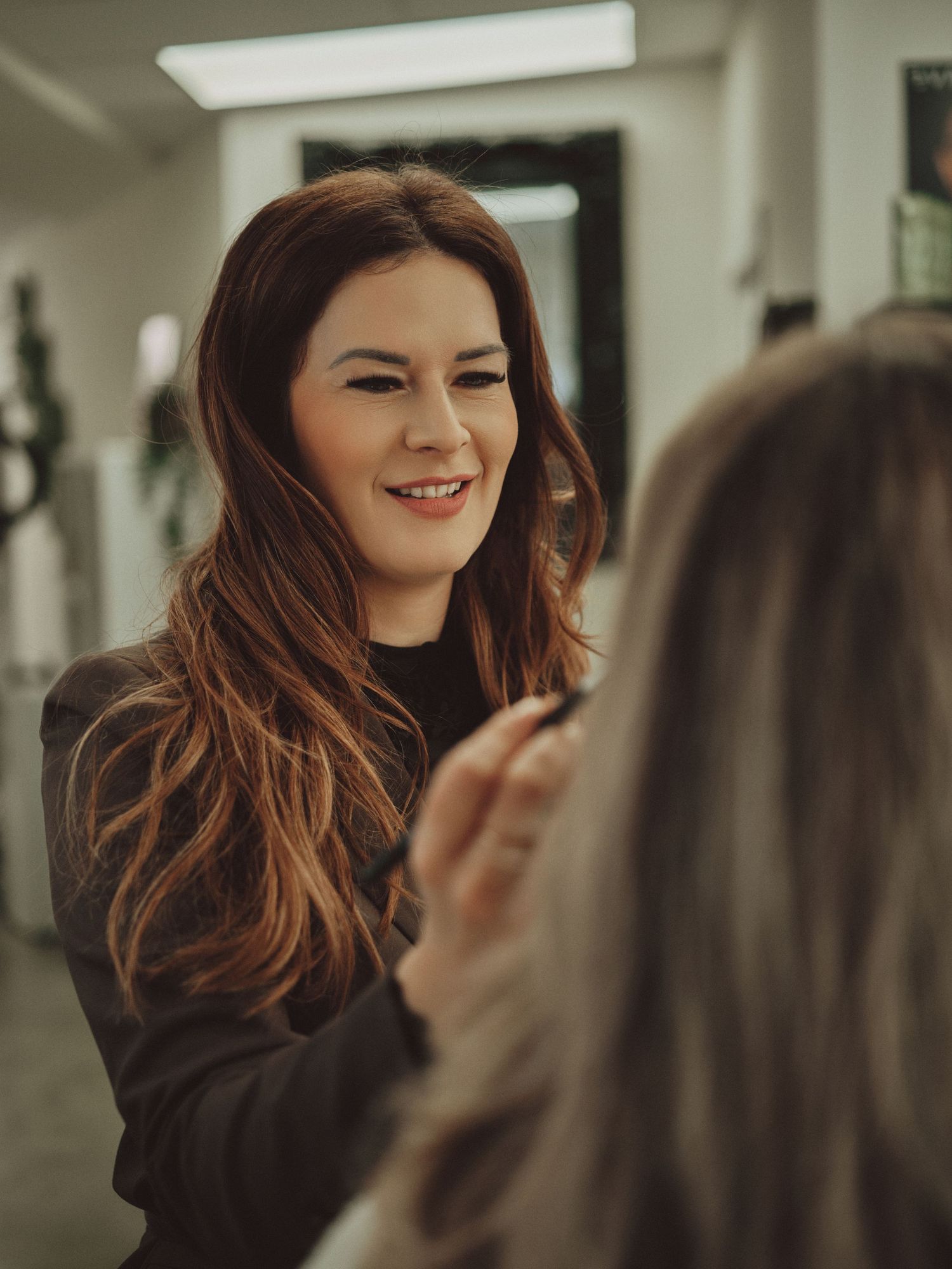 A smiling makeup artist with long auburn hair applies makeup to a client in a bright salon studio.