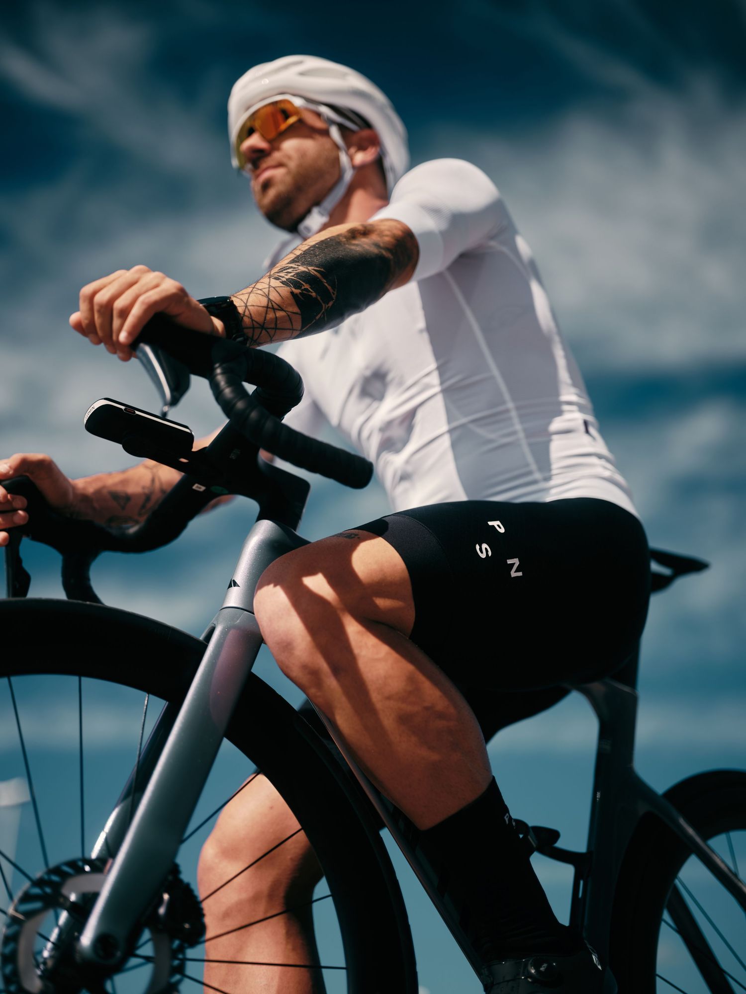 Tattooed male cyclist in white jersey and black shorts poses with a carbon road bike against a blue sky.