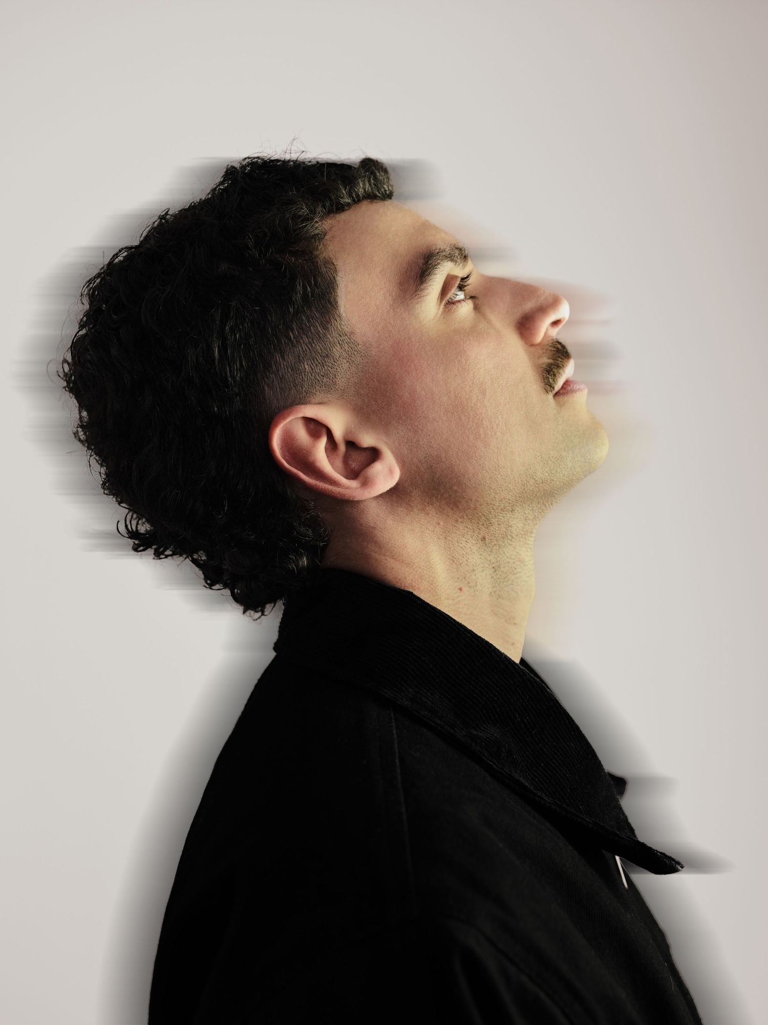 Side profile of man with curly dark hair in black jacket with motion blur effect on white background.