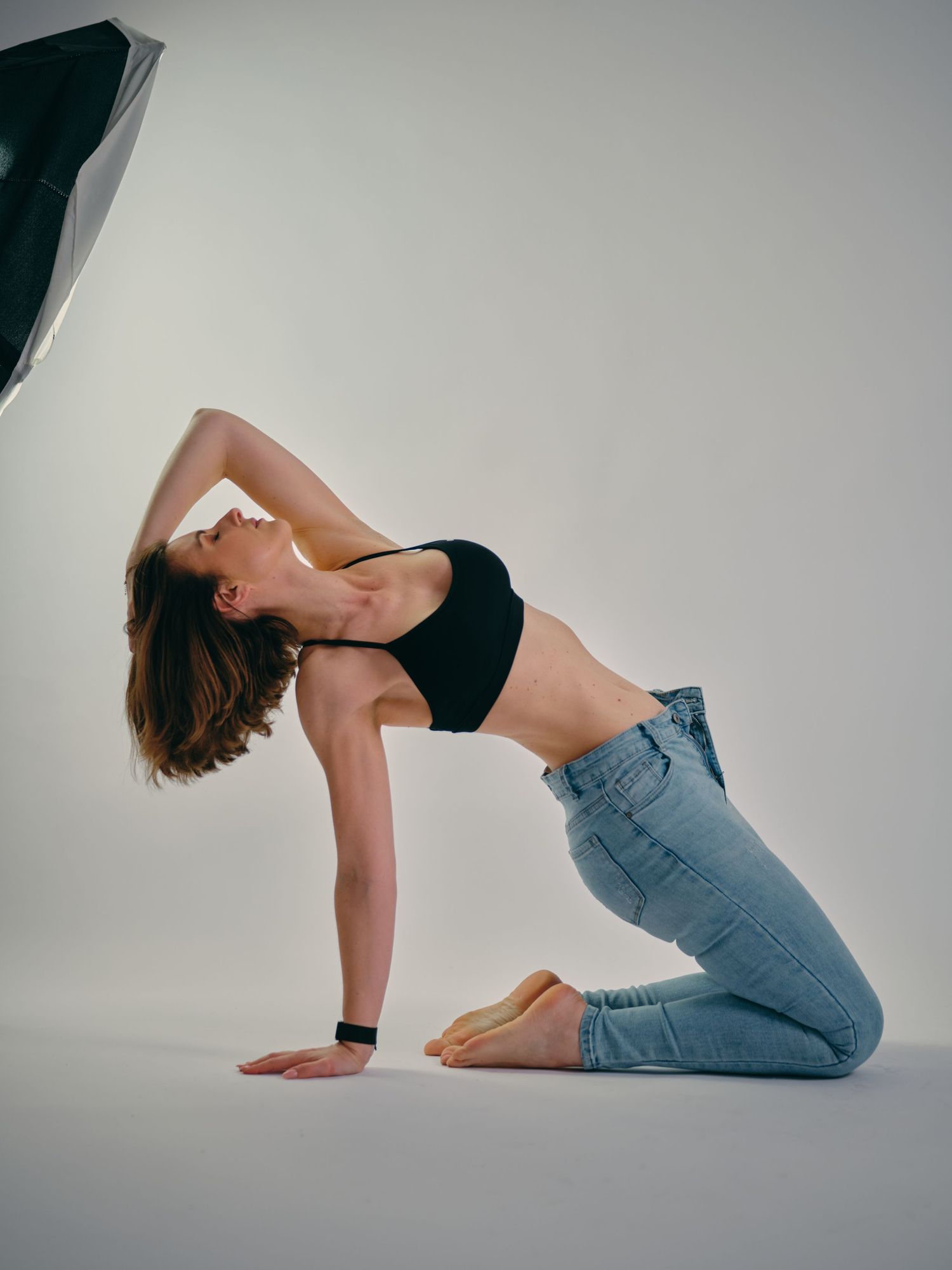 Woman in black crop top and jeans performing a yoga backbend pose on knees in a professional photo studio.