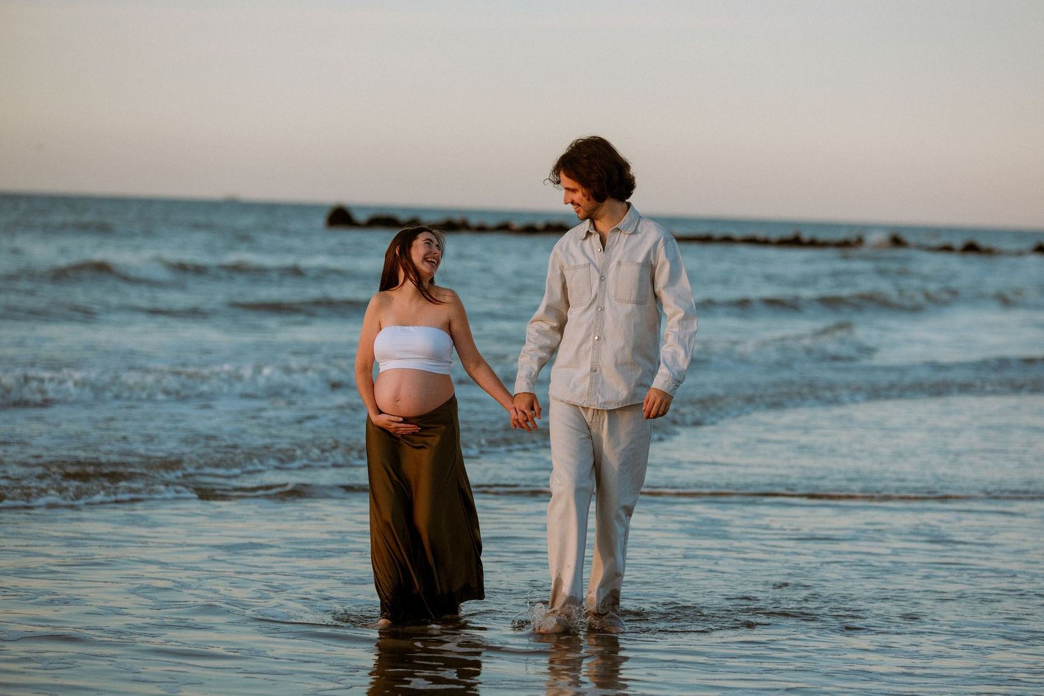 A couple stands together holding hands in shallow ocean water at sunset, creating a romantic beach scene.