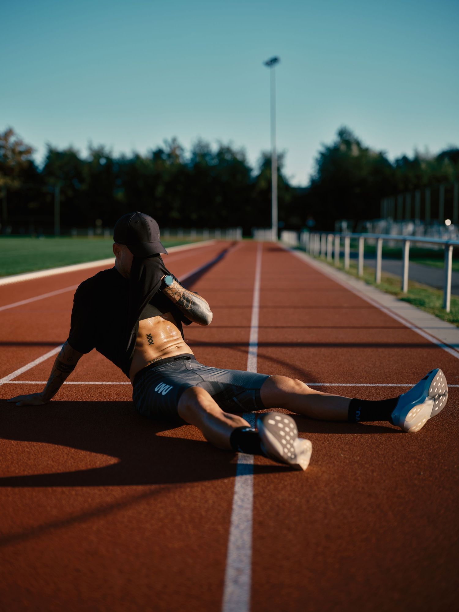 Athlete stretching on a red running track outdoors, wearing shorts and a black jacket, with trees in the background.