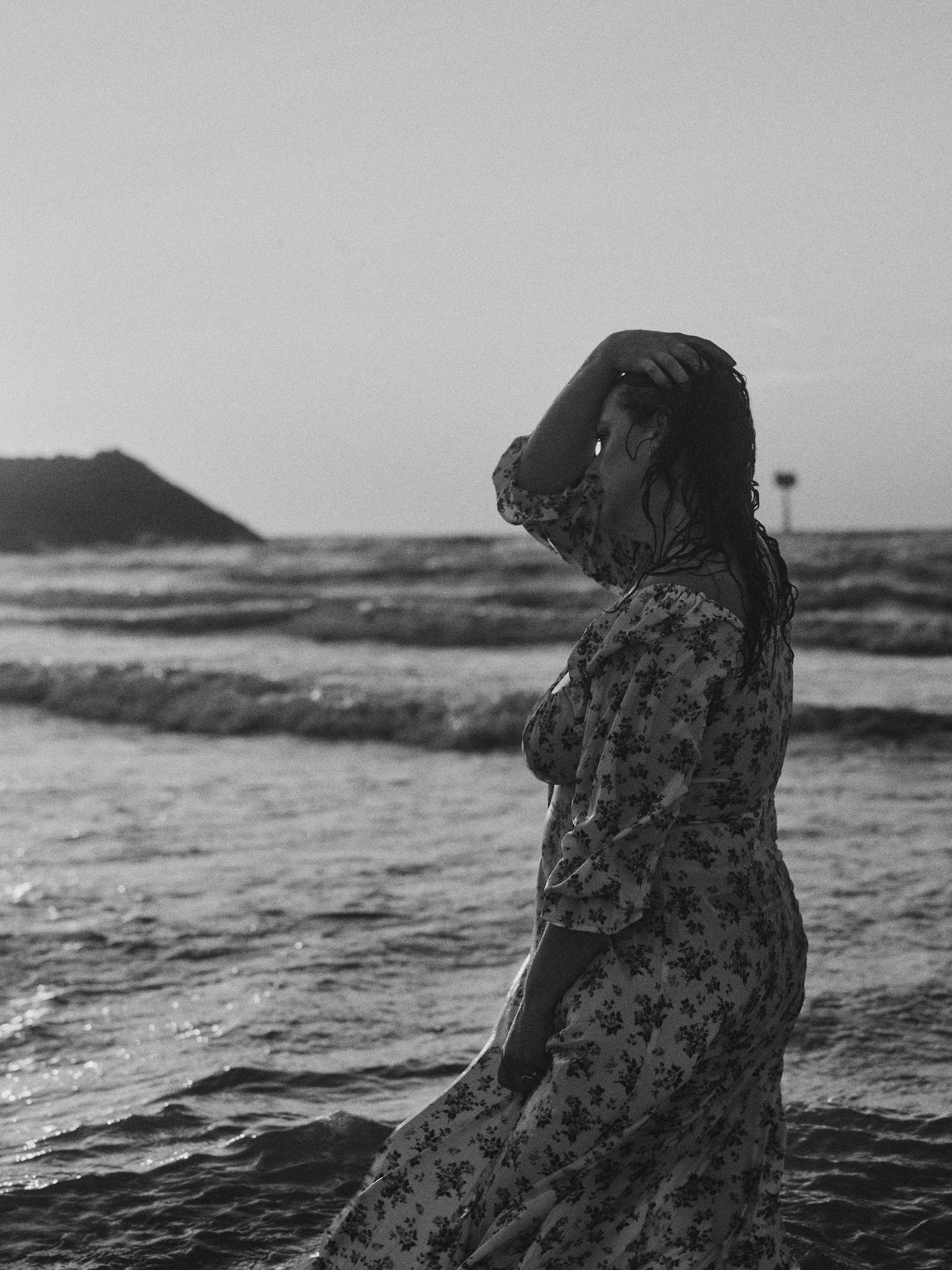 Black and white photo of a woman in a floral dress standing on a windy beach with ocean waves behind her.