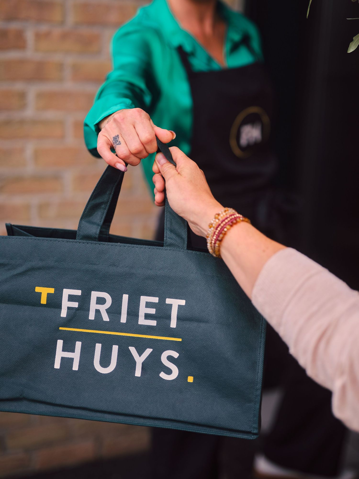 A branded T Friet Huys dark tote bag being handed between two people, one wearing a green shirt and black apron.