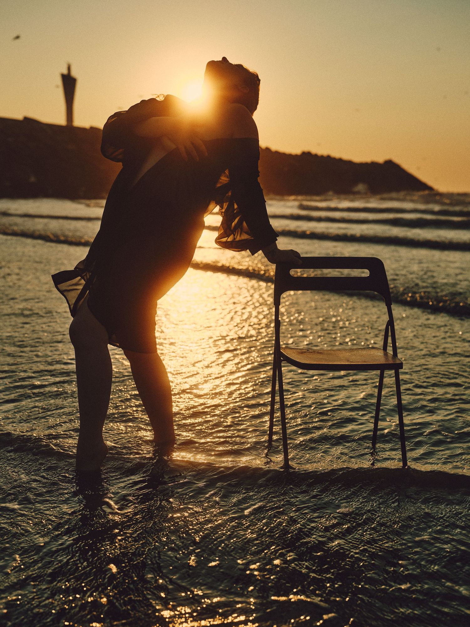 Silhouette of a person dancing on a beach at golden sunset with a chair in shallow water.