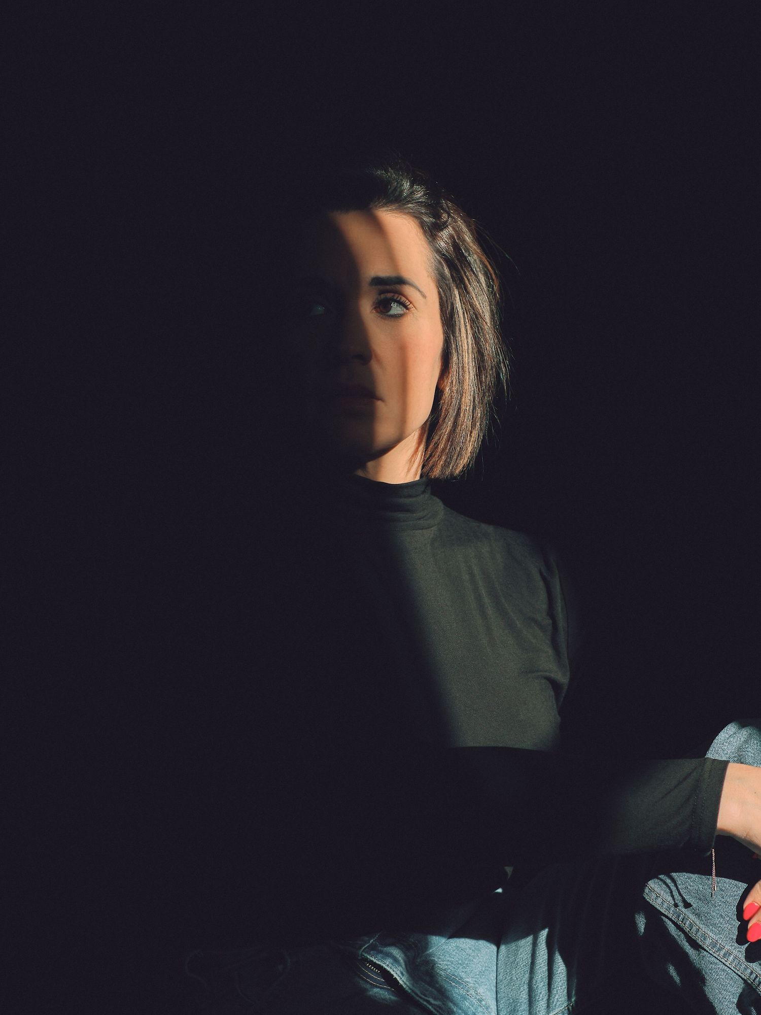 A person with shoulder-length hair wearing a black turtleneck, dramatically lit against a dark background.