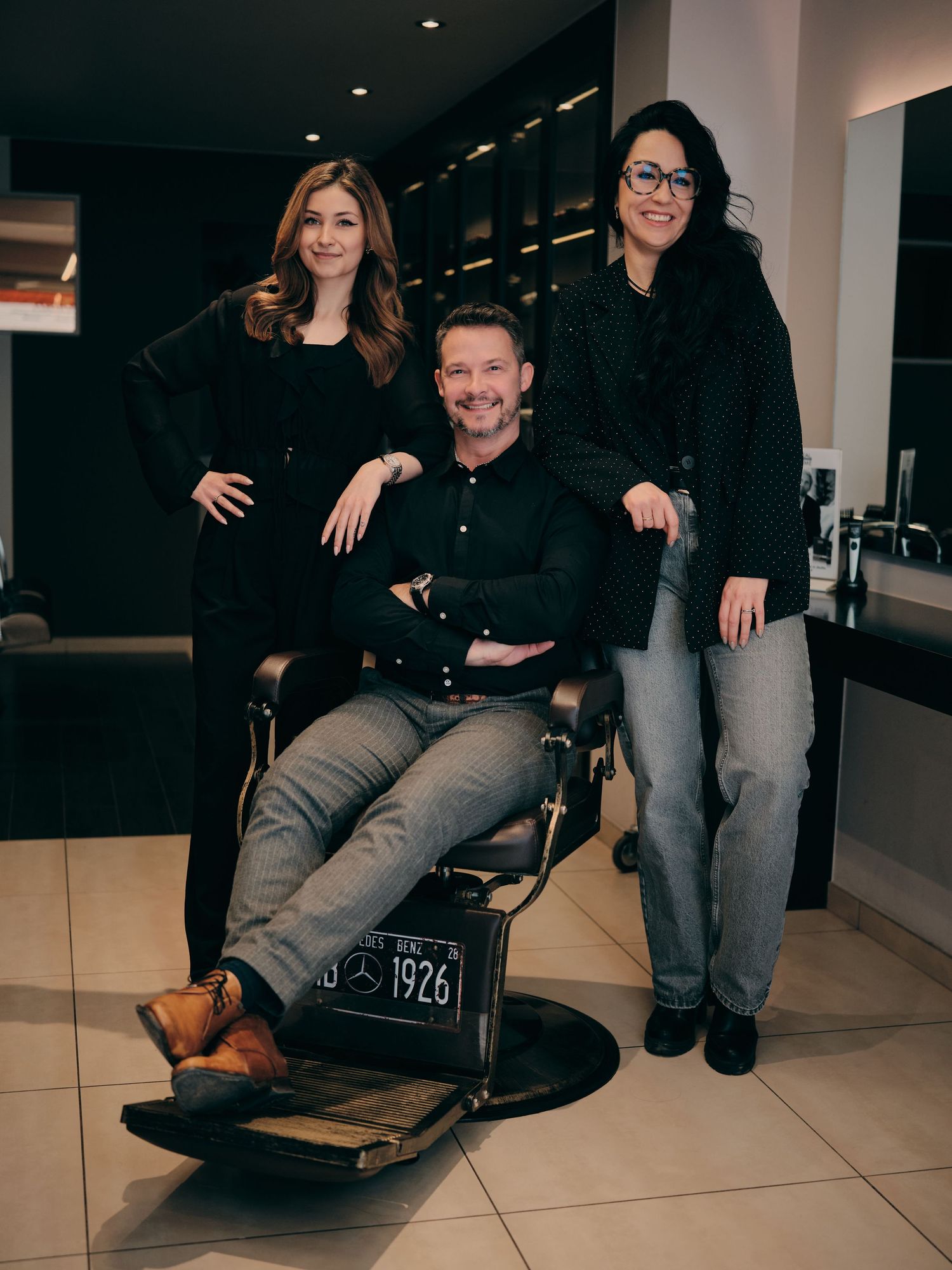 Three salon professionals pose in a modern barbershop, one seated in a vintage 1926 barber chair, all dressed in black.