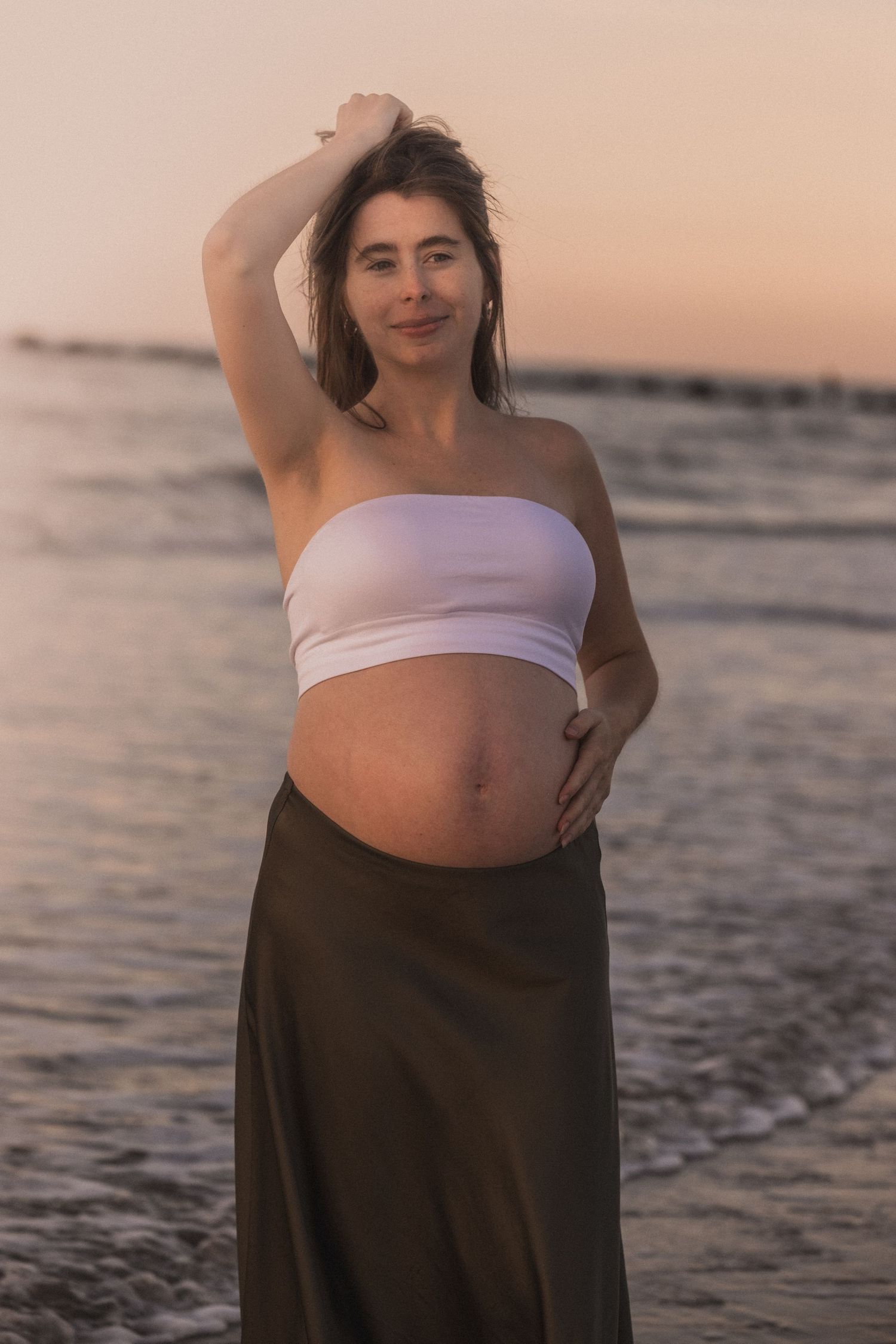Maternity photo shoot at sunset on the beach showing a pregnant woman in white top and dark skirt.