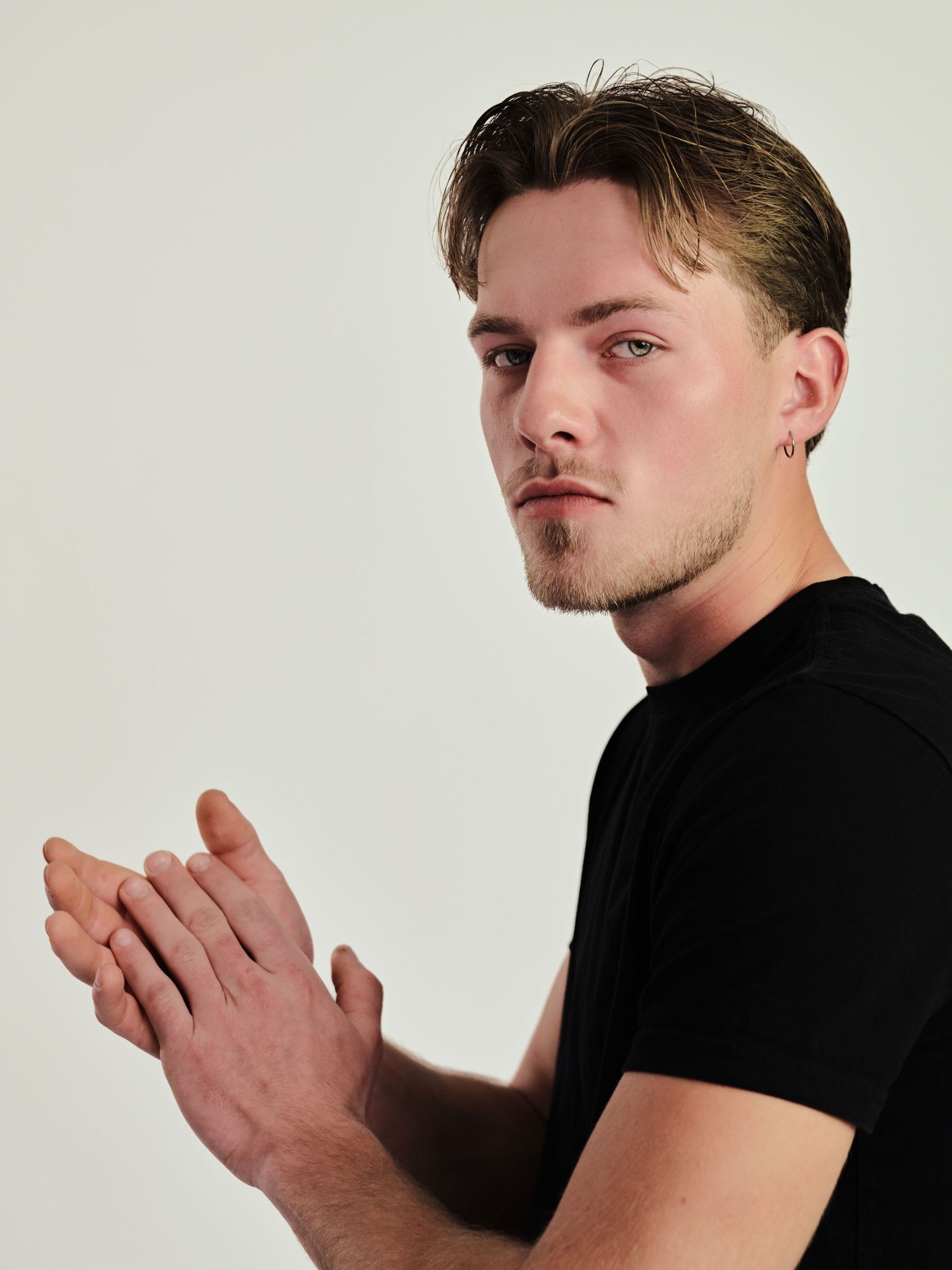 Young man in black t-shirt with ear piercing clapping hands, looking over shoulder against white background.