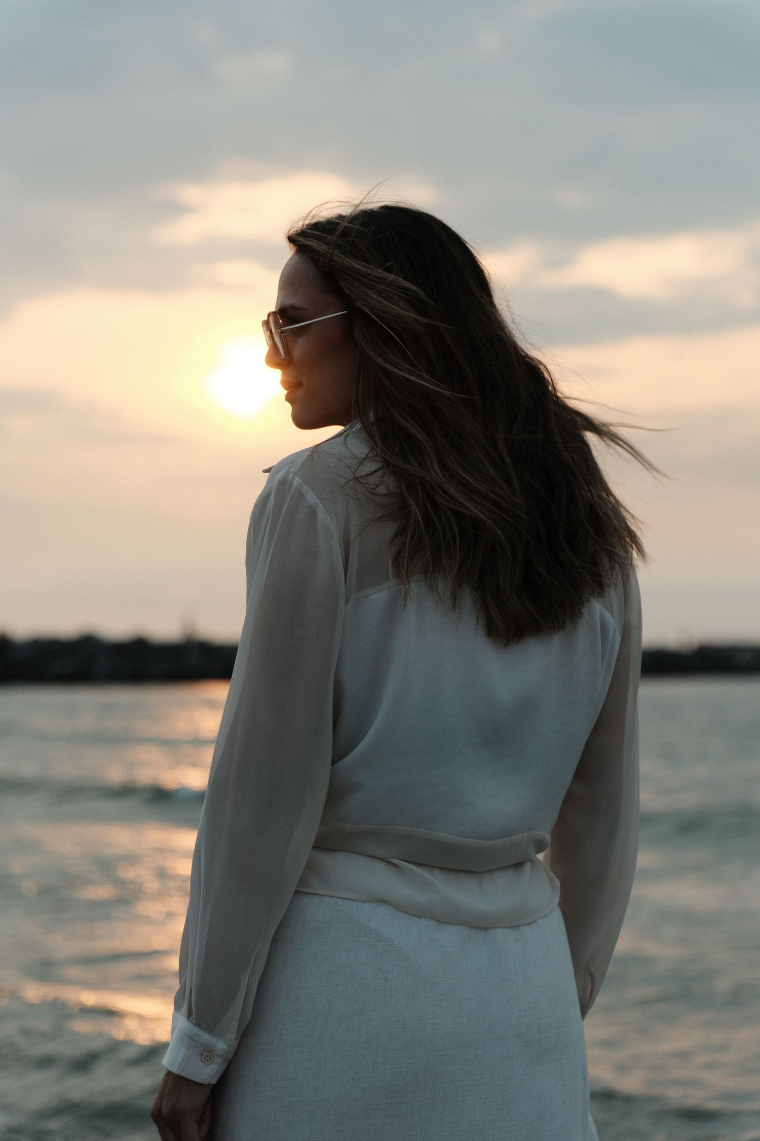 Woman in white dress with sunglasses gazing at a serene sunset over the water, hair blowing in the breeze.