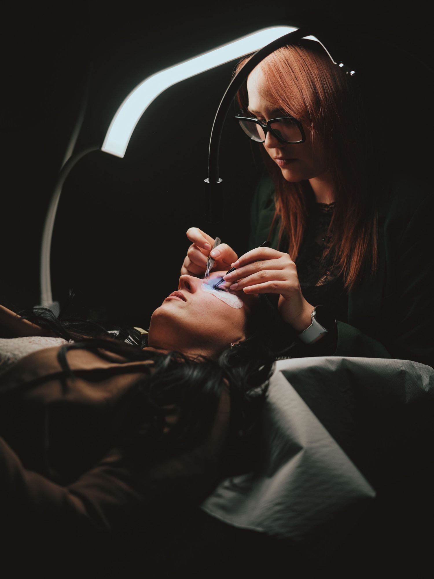 A beauty technician with glasses applies lash extensions to a client under bright salon lighting in a dark studio.