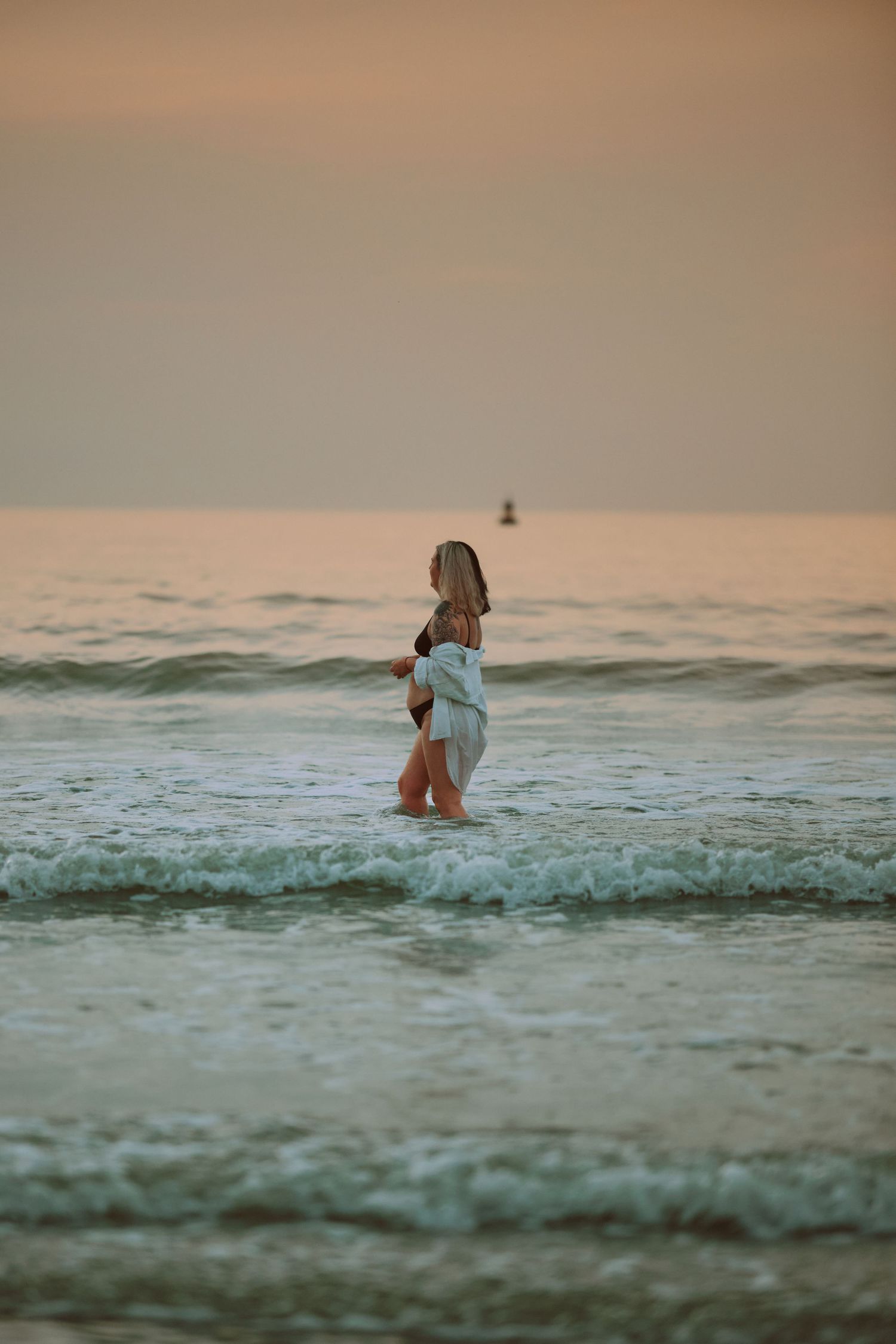 Woman standing in ocean waves at sunset wearing a white outfit, with calm sea and golden sky in the background.