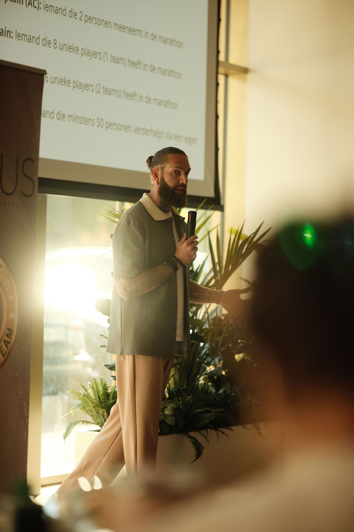 Bearded man in sage green outfit presenting at a sunlit conference, holding a microphone with plants in background.