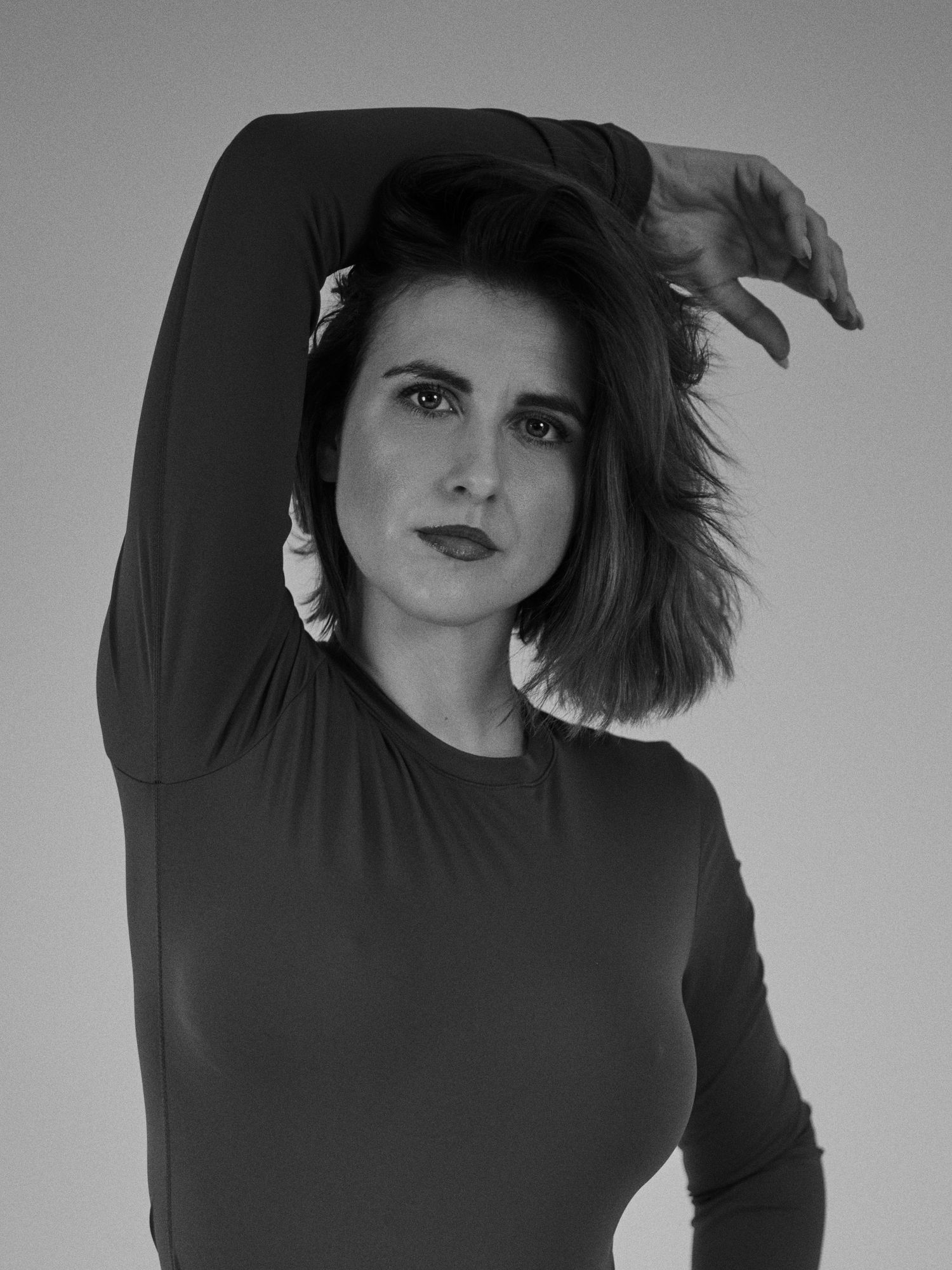 Black and white portrait of a woman in a dark long-sleeve top posing with arm raised gracefully overhead.