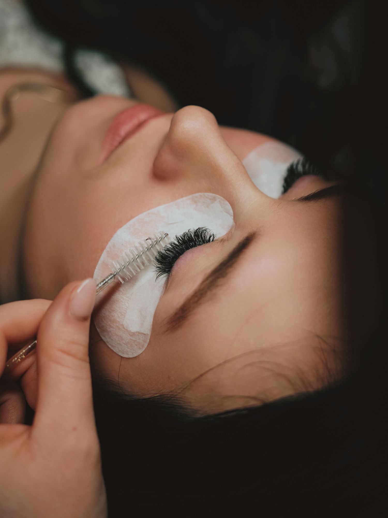 A beauty technician applies eyelash extensions to a woman lying down with white under-eye pads during a lash treatment.
