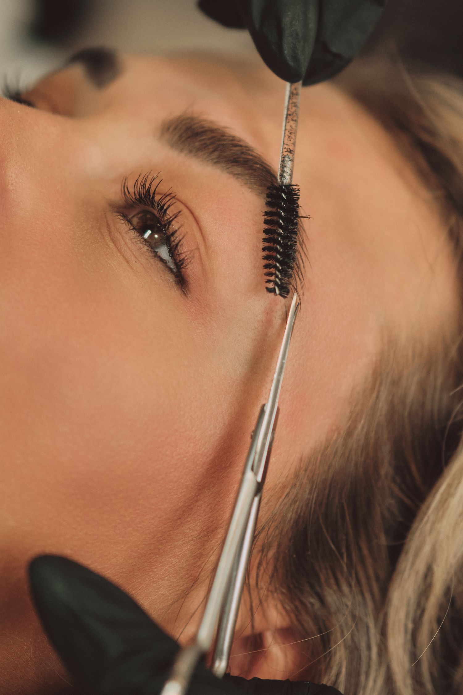 Esthetician grooming womans eyebrows with spoolie brush and scissors in professional beauty salon.