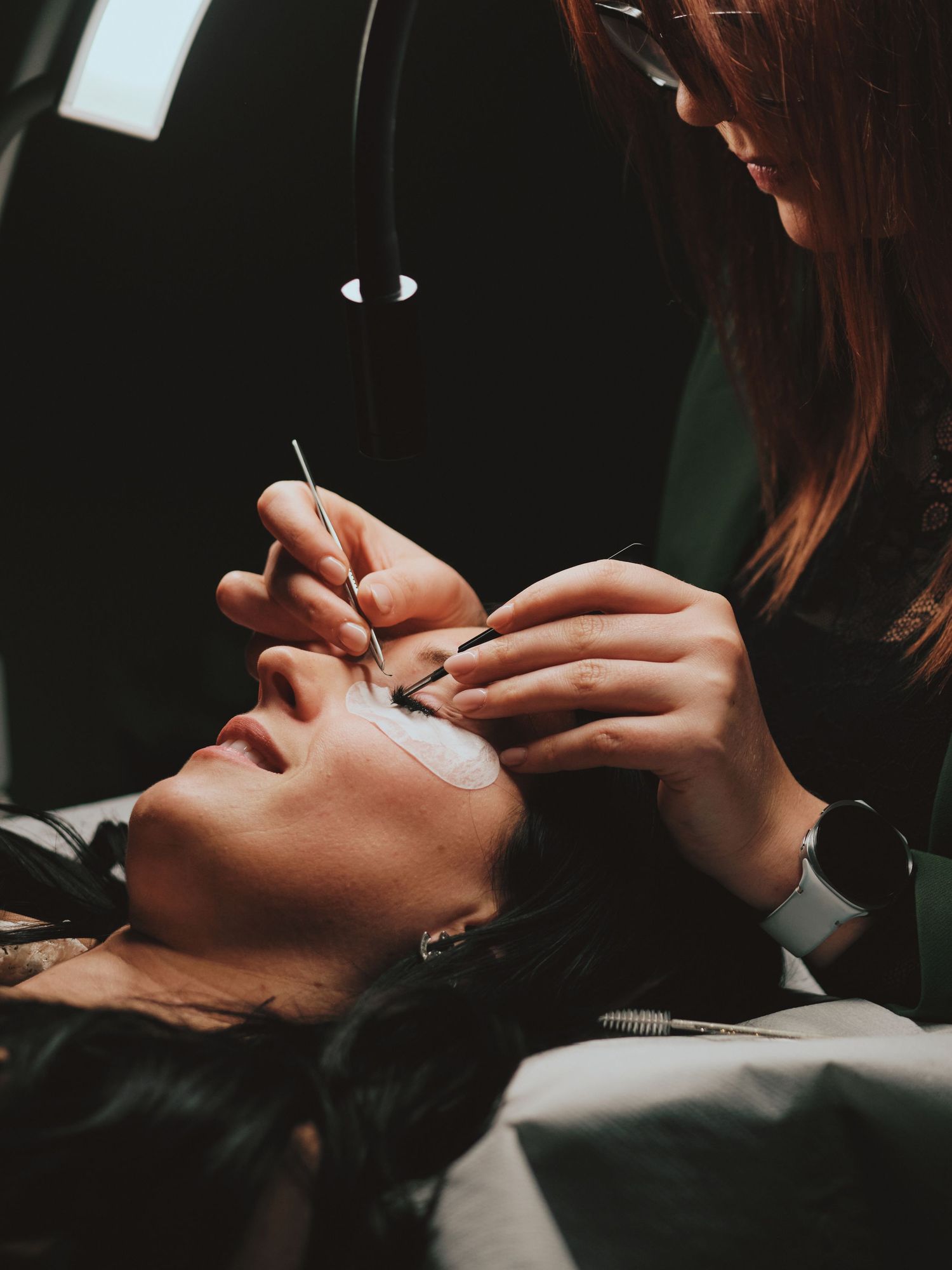 Esthetician applying eyelash extensions to a client lying down, using precision tweezers under a bright salon lamp.