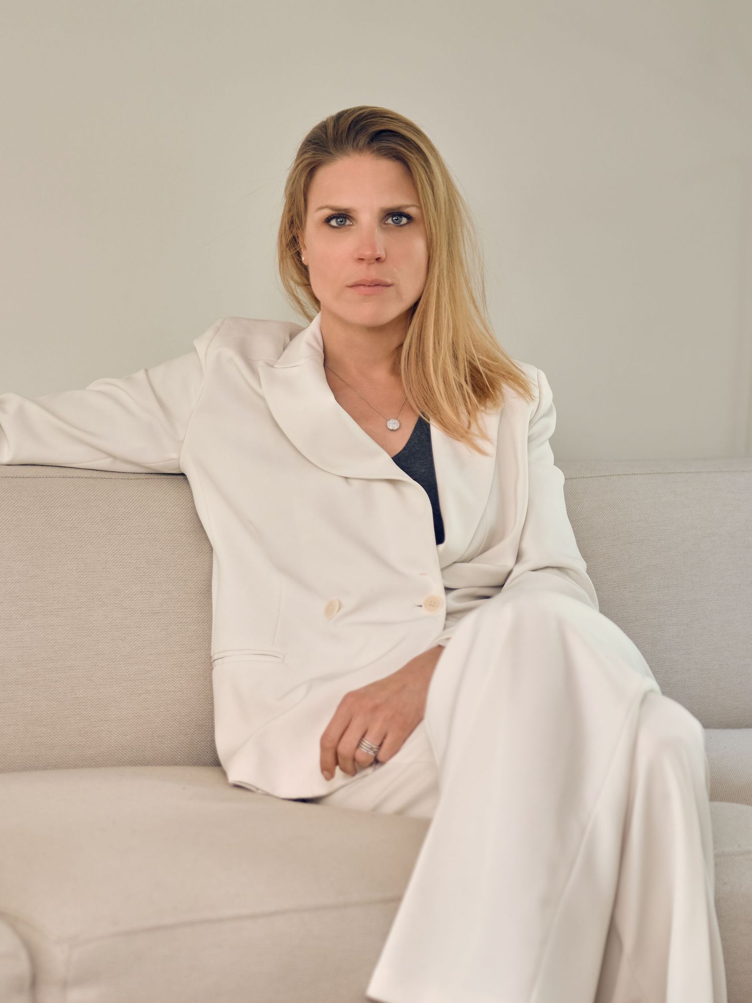 Woman in white blazer and pants sitting on a beige sofa with a neutral background.