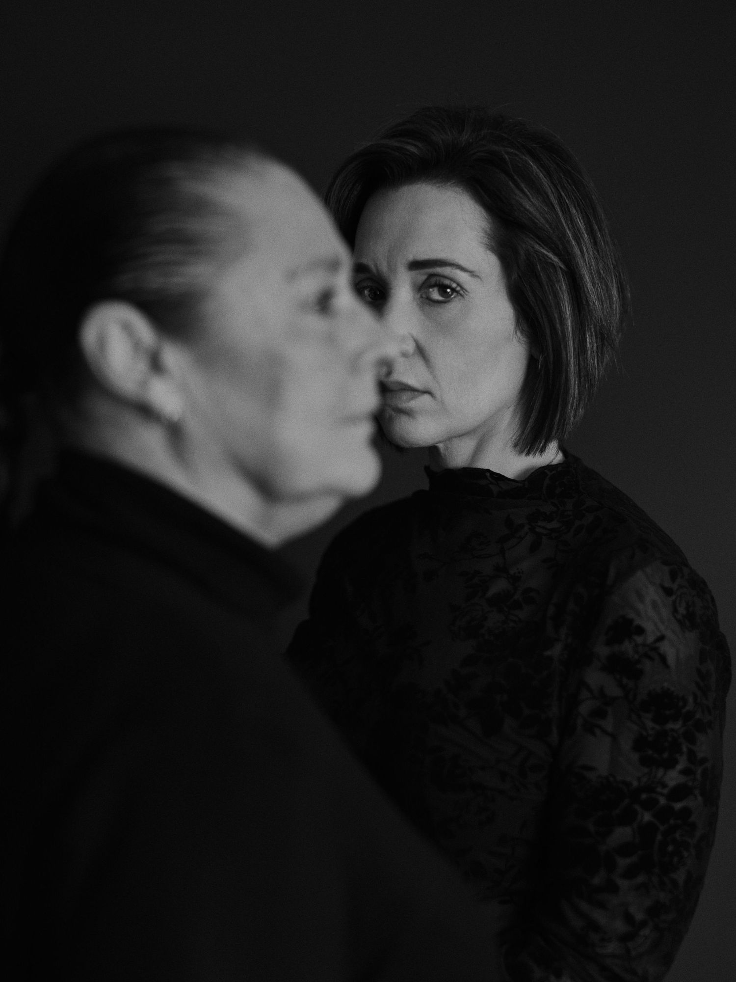 Black and white portrait of two women in dark clothing against a dark background, one in focus, one blurred.