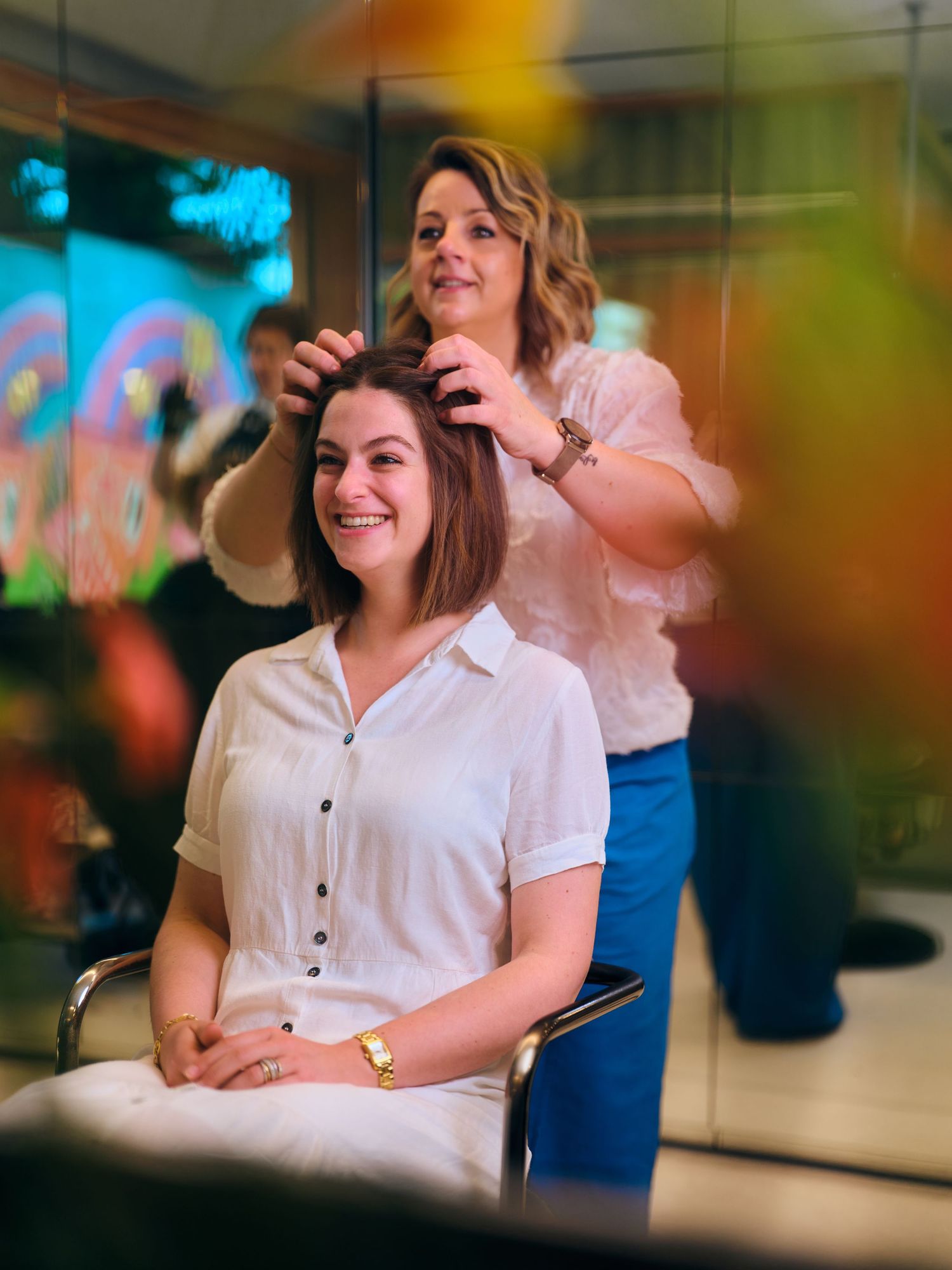 A smiling woman gets her hair styled by a hairdresser in a modern salon with colorful lighting.