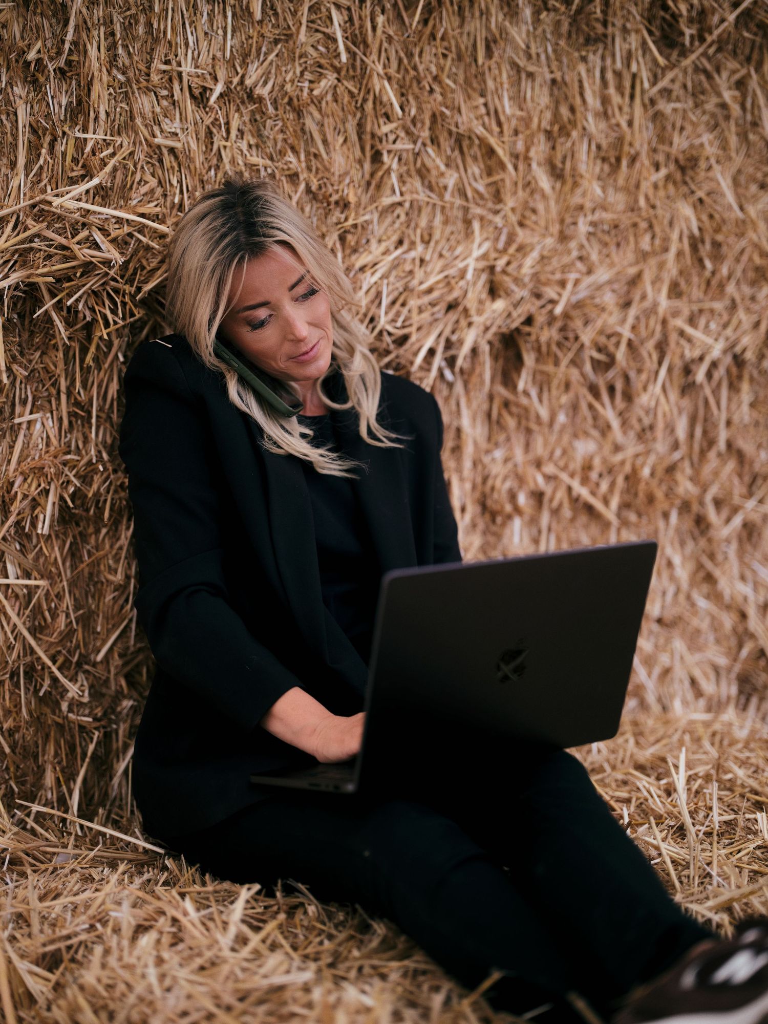 Woman in black outfit working on laptop while sitting against hay bales in a rustic farm setting.