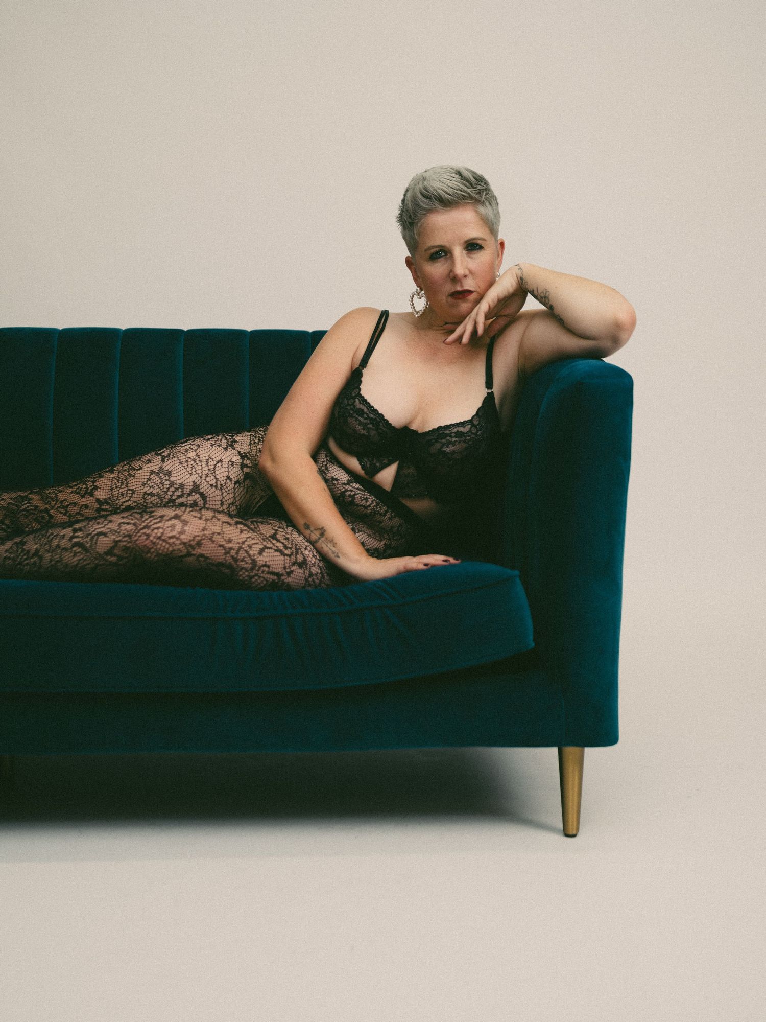A person in a black dress reclines on a dark blue velvet couch against a beige wall in a moody photoshoot series.