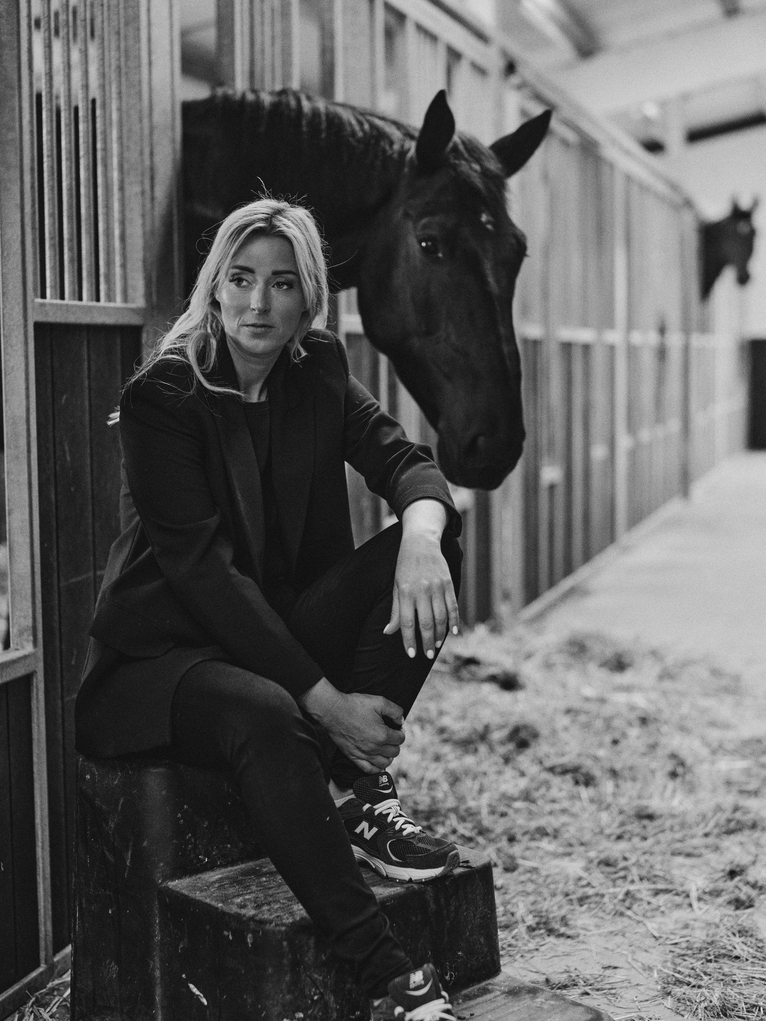 Black and white photo of a blonde woman sitting in a stable beside a dark horse in New Balance sneakers.