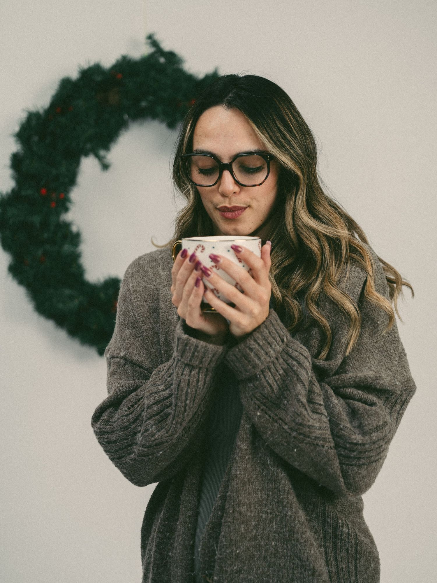 A person in a cozy gray sweater holds a warm mug near a dark wreath in a minimalist winter scene.