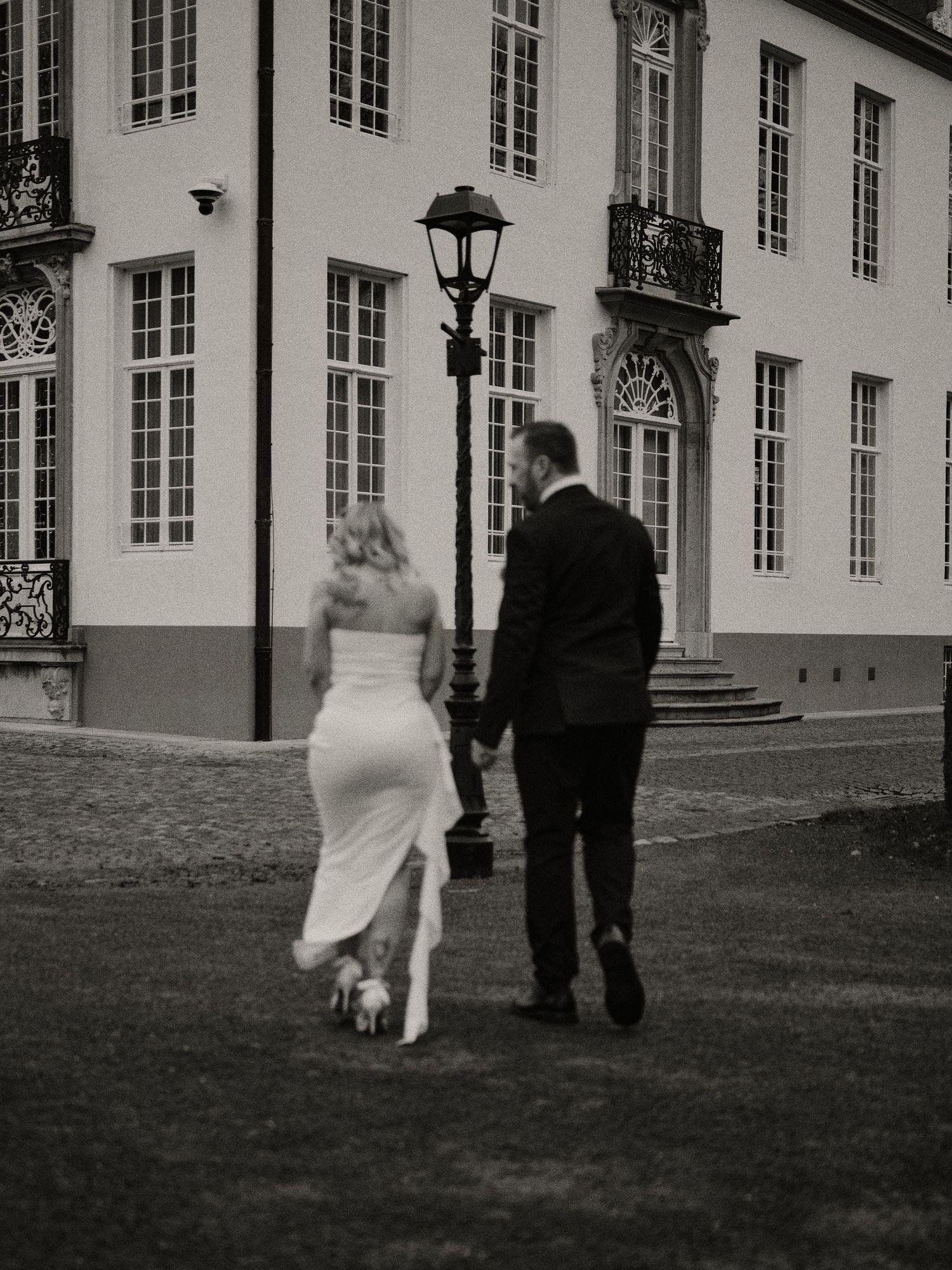 A couple walks together on the lawn of an elegant historic building with white walls and tall windows in black and white.