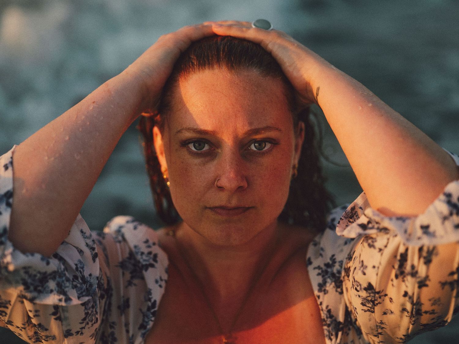 Woman with wet hair holds hands on head, gazing at camera in warm golden sunset light near water.