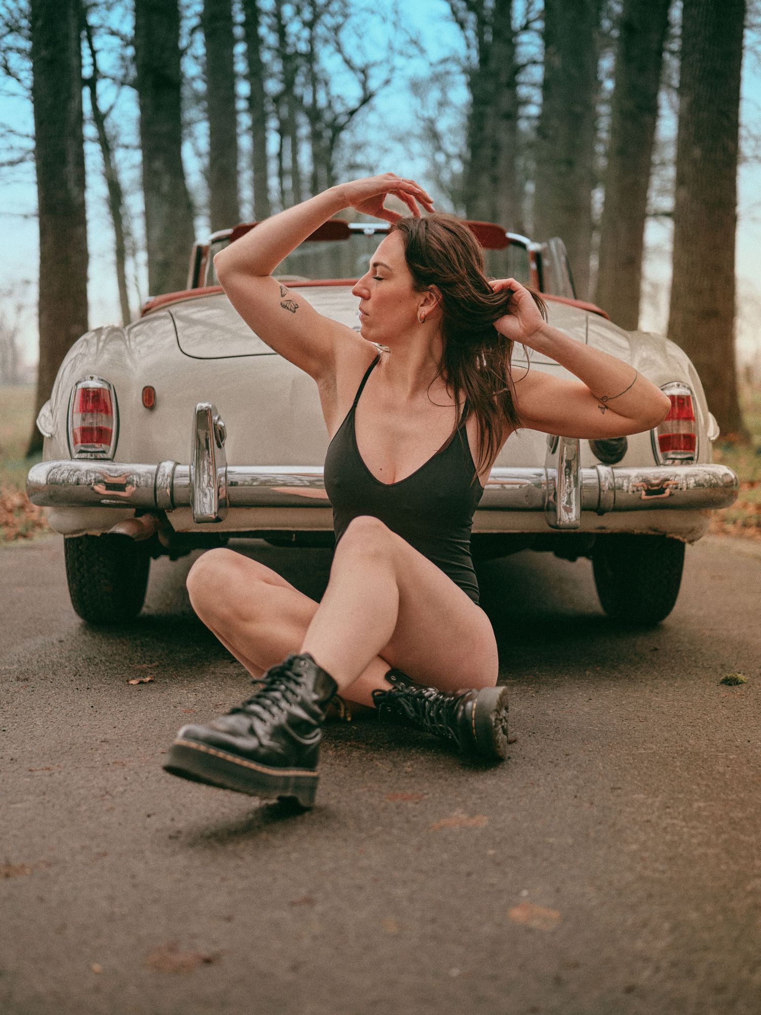 A woman in a black bodysuit and Dr. Martens boots sits on a road in front of a vintage cream-colored car.