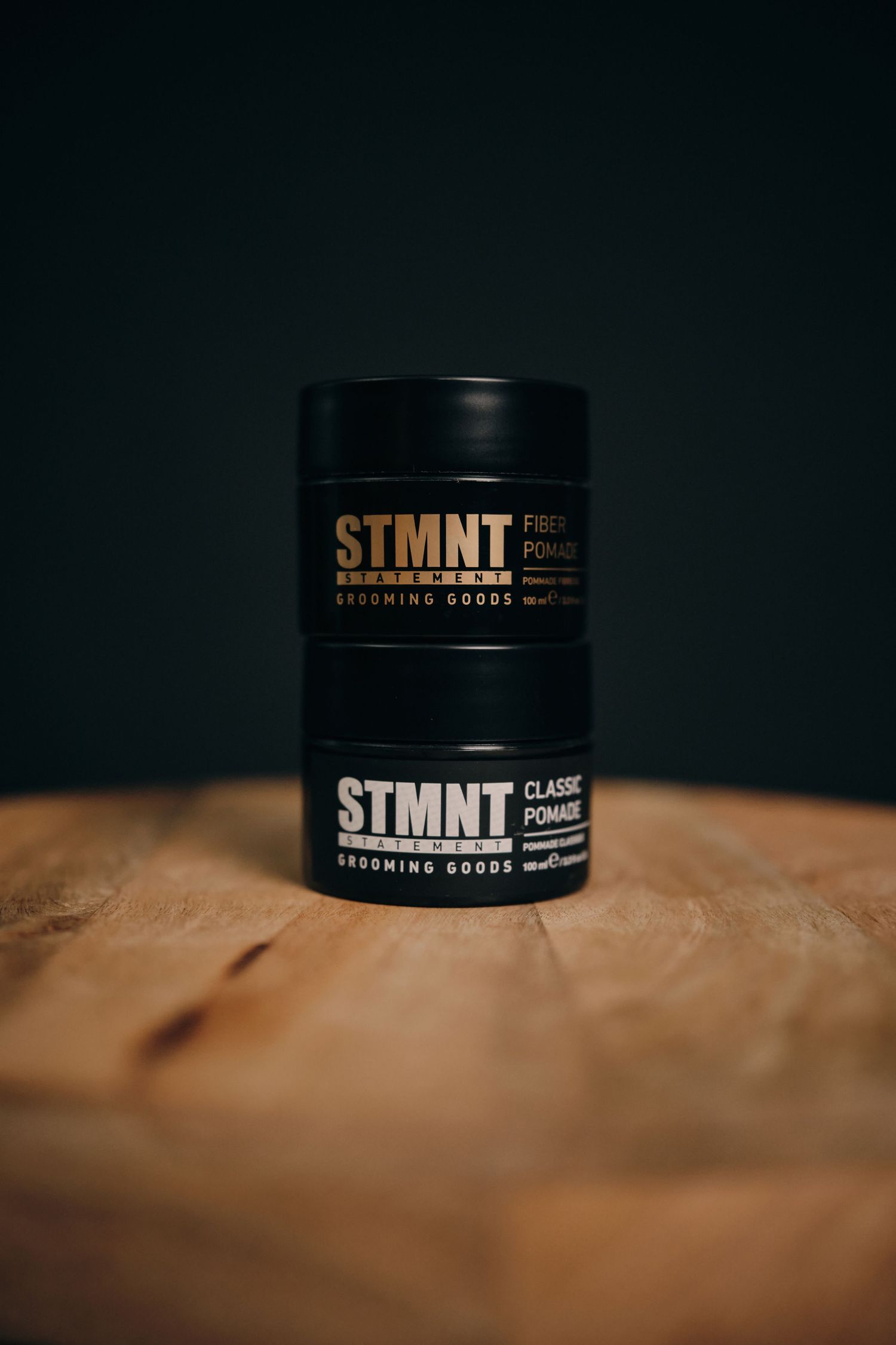 Black STMNT grooming products jar displayed on textured wooden surface with dramatic lighting and shallow depth of field.
