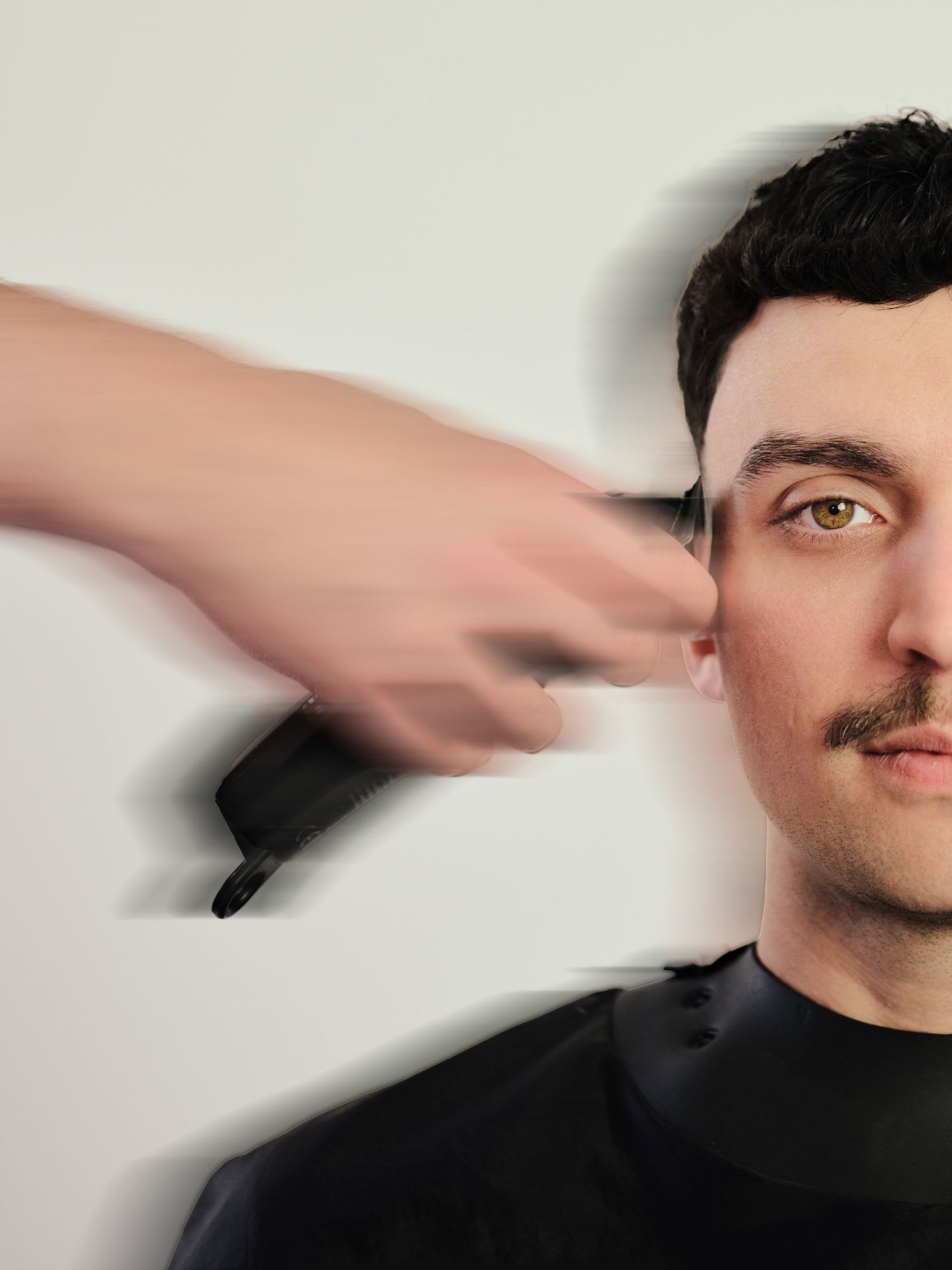 A barber uses clippers to trim the sides of a young man with a mustache wearing a black cape.