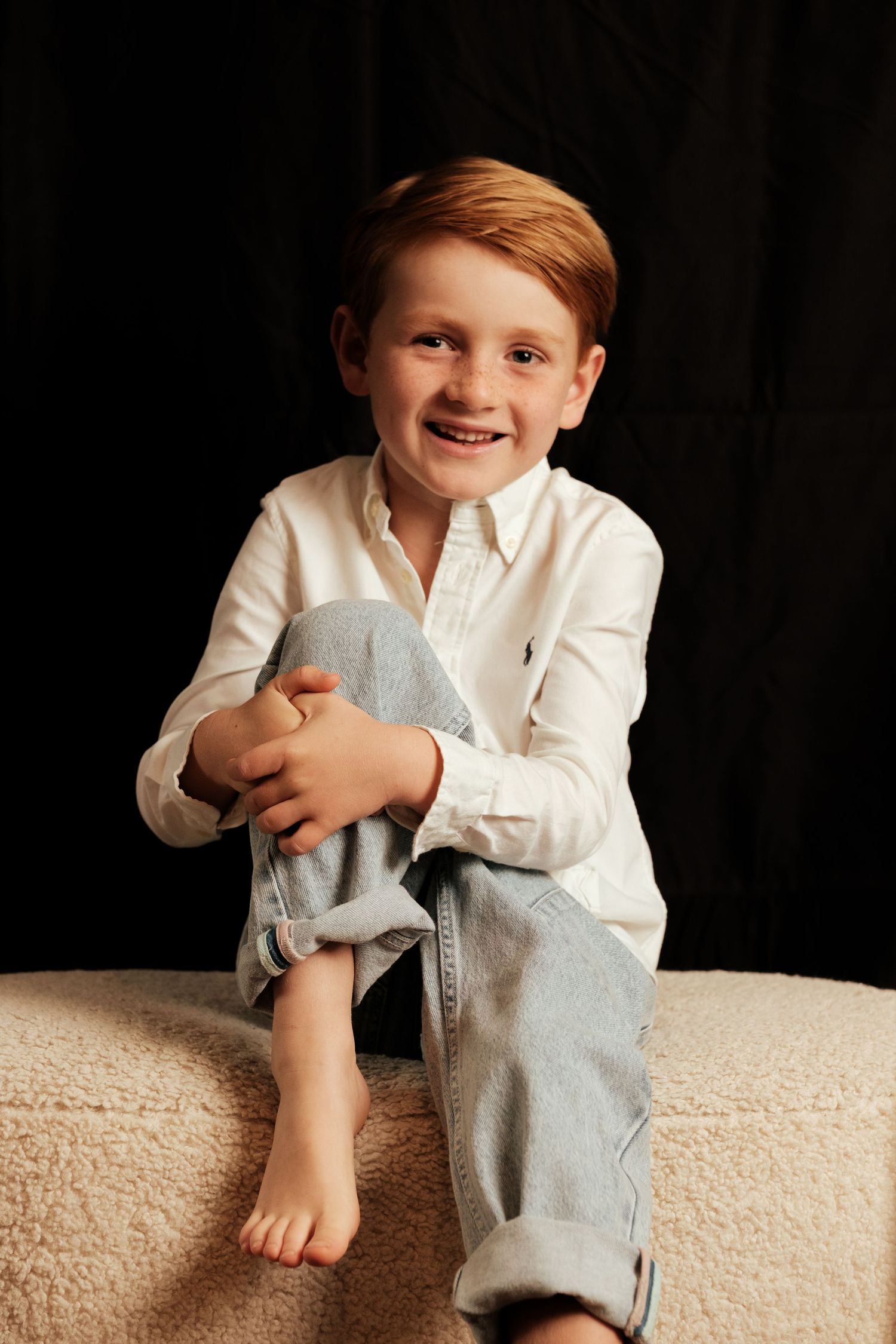 Child in white button-down shirt and gray pants sits casually on beige ottoman against black background.