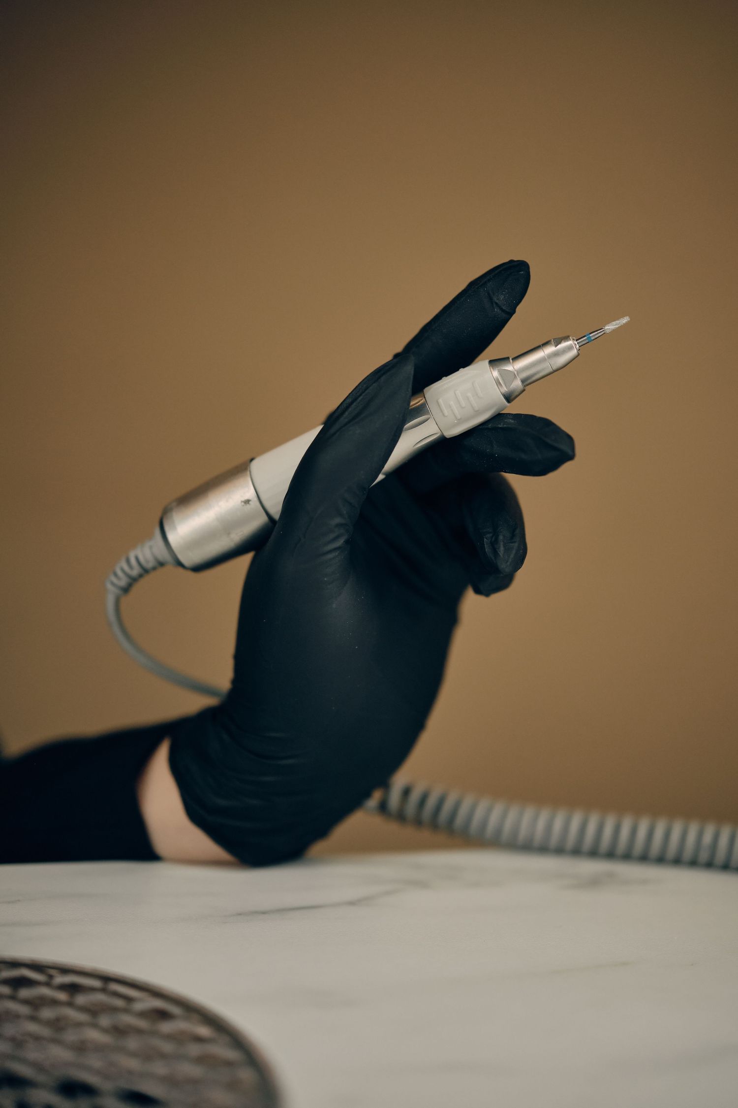 A gloved hand holds an electric nail drill tool against a neutral beige background in a nail salon setting.