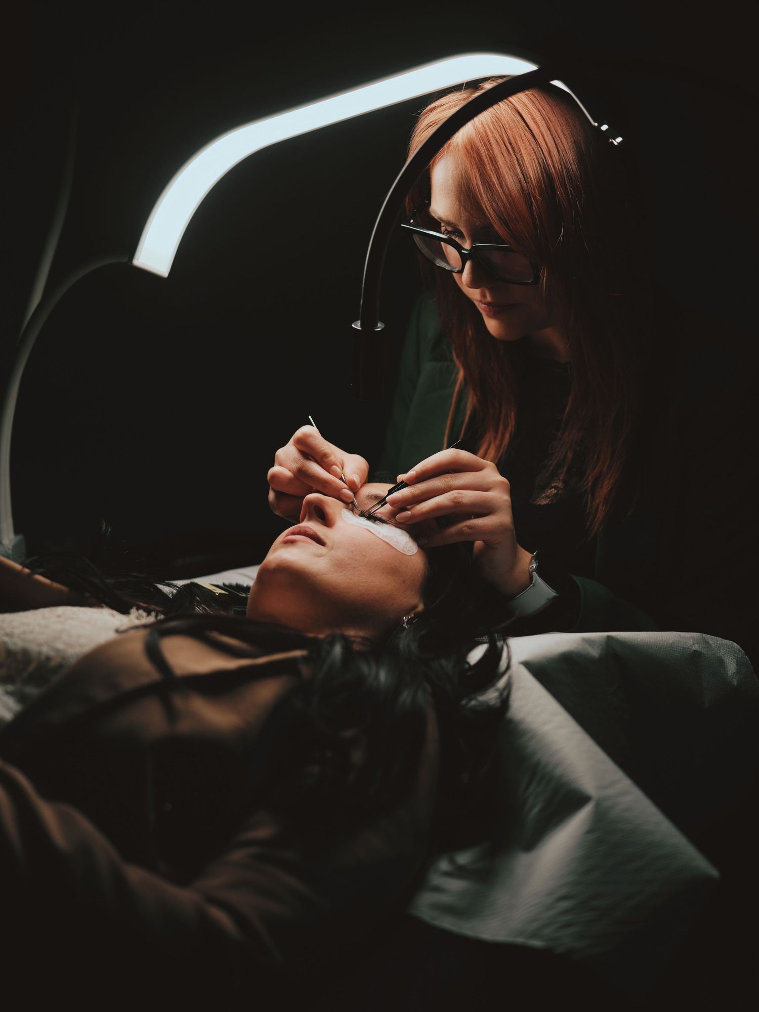 A beauty technician with red hair applies eyelash extensions to a client under a bright lamp in a dark salon.