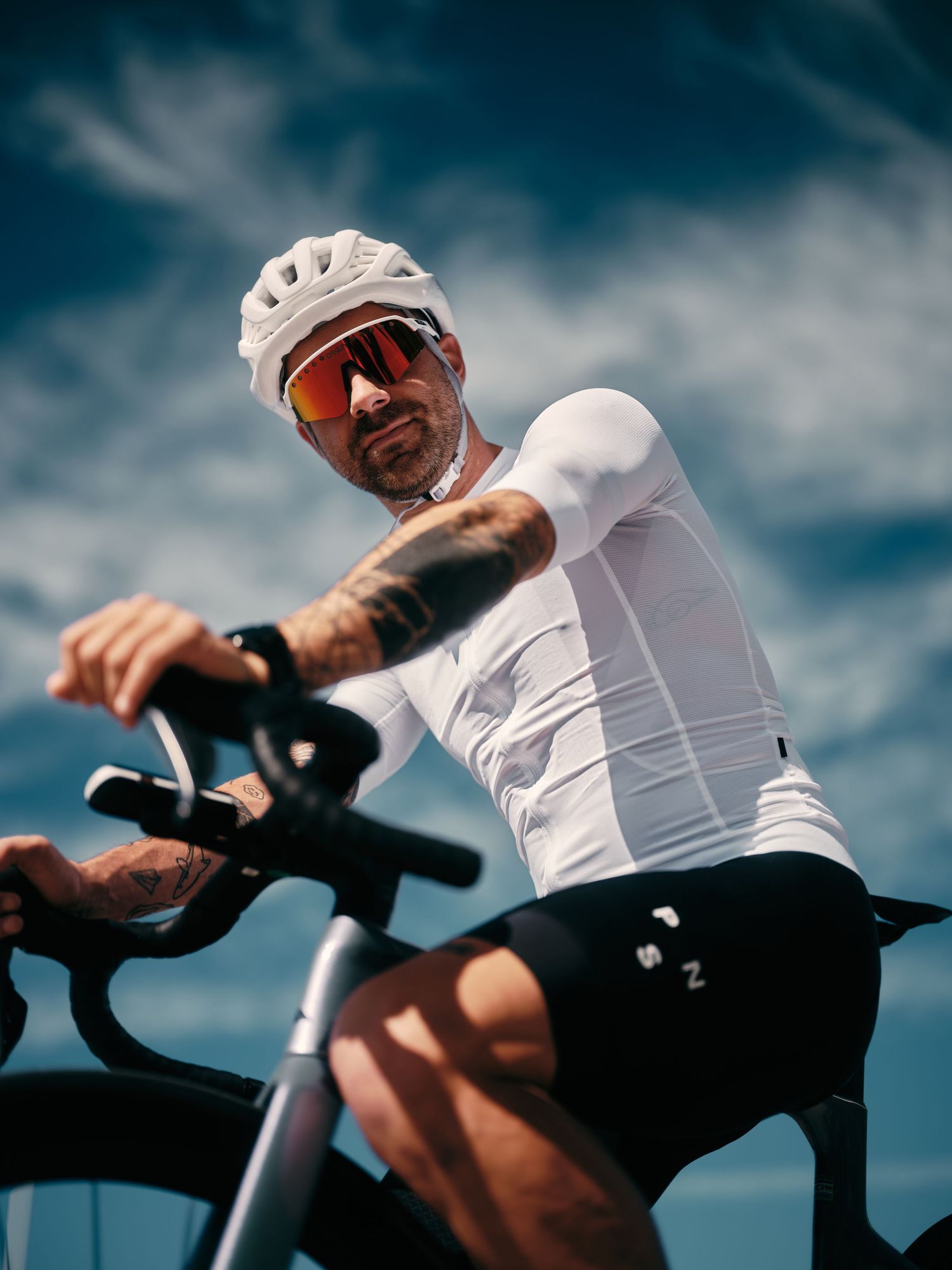 Tattooed male cyclist in white jersey and helmet rides a road bike against a dramatic blue sky.