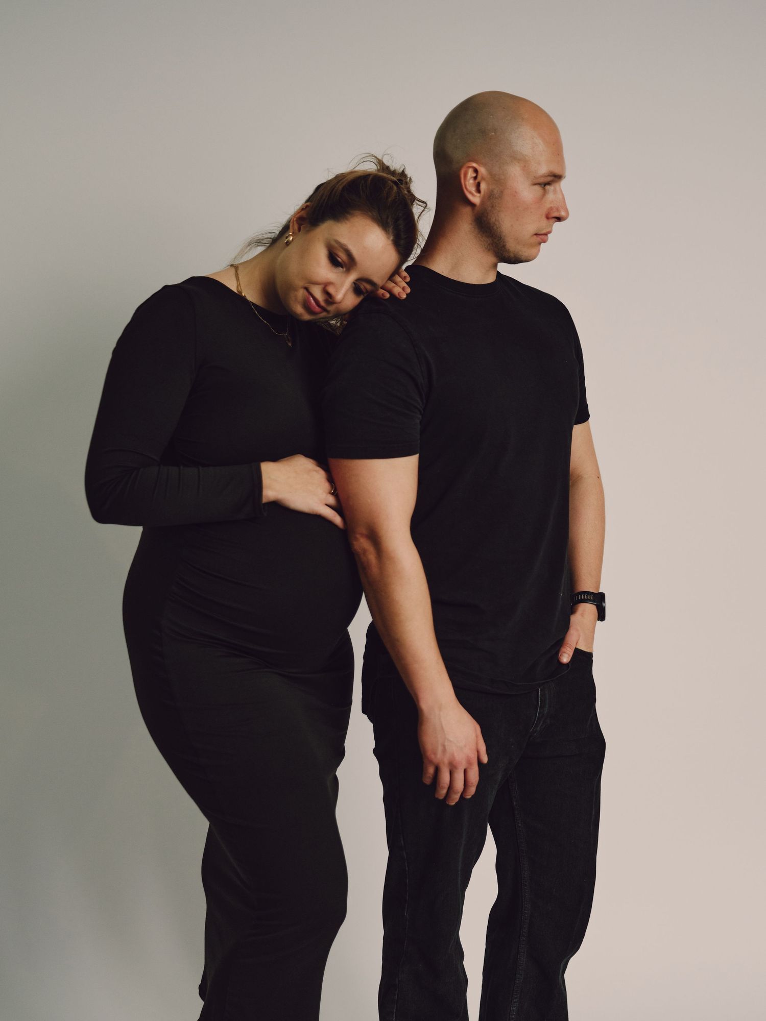 Two people in black clothing embrace in an intimate dance-like pose against a light gray studio background.
