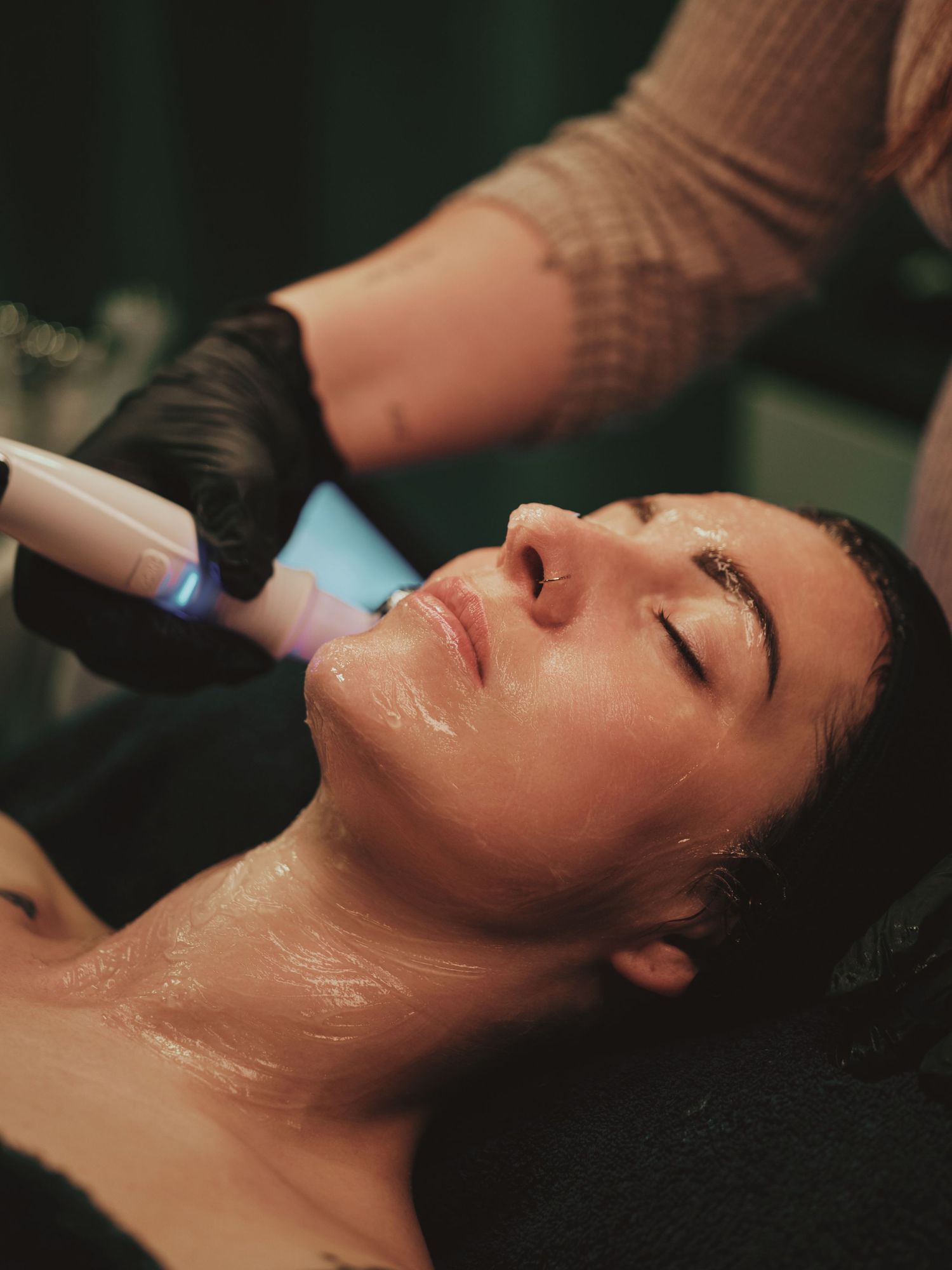 A woman receives a hydrafacial treatment at a spa, with a glowing device applied to her face by a gloved esthetician.